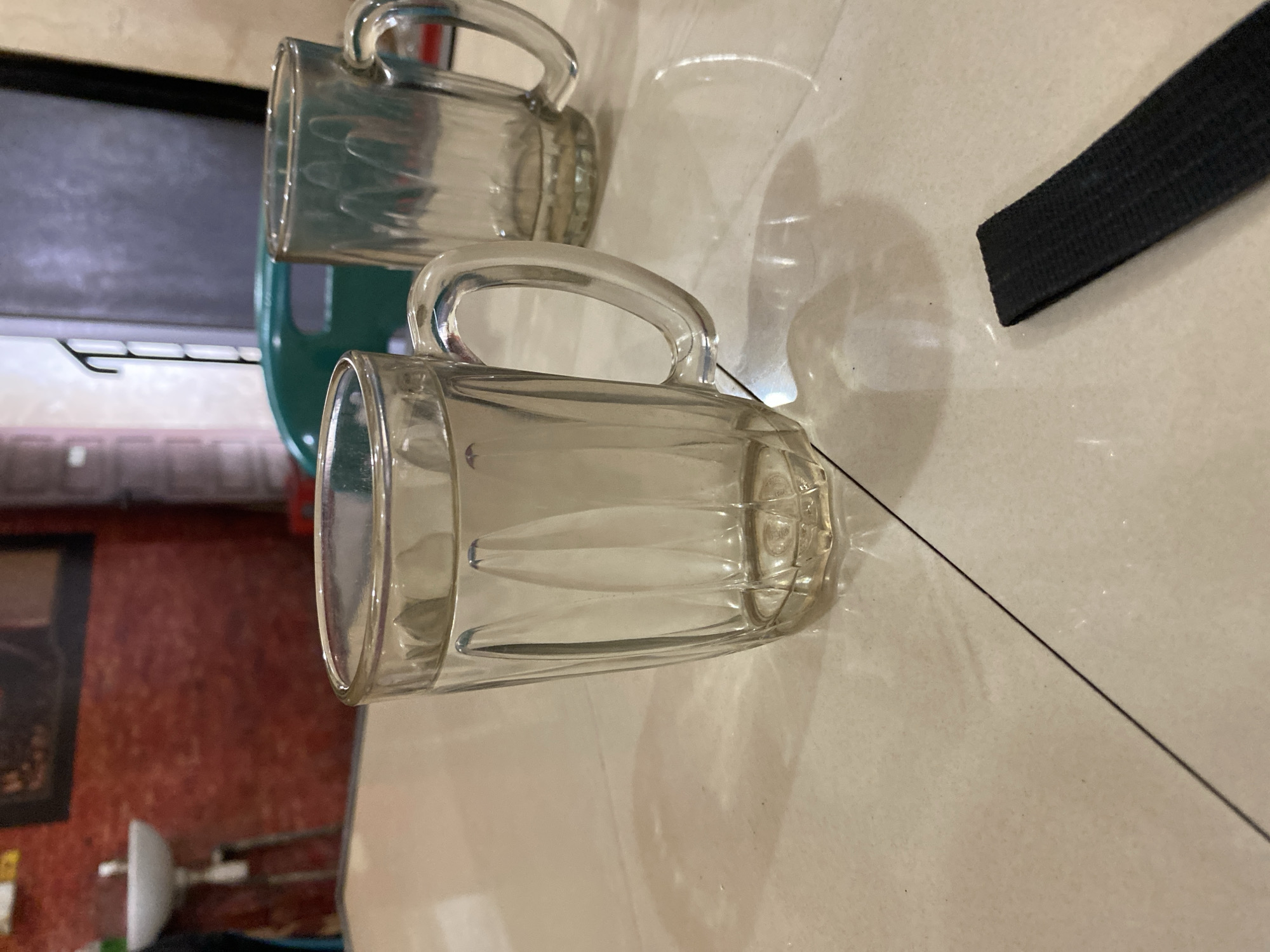The image shows two clear glass mugs with handles. One mug is positioned in the foreground, slightly to the right, and is empty. The other mug is behind it, slightly to the left, and appears to be empty as well. Both mugs have a ridged texture on their sides. The mugs are placed on a light-colored tiled surface. In the background, to the left, a dark green object is visible, possibly a container or another mug. Further in the background, a black strap or band is seen, angled across the surface. To the far left, a framed picture with a dark border is visible on a red textured wall, and what seems to be a white object with metal supports is present on a lower surface. The lighting suggests it is daytime, with shadows cast on the tiled floor. The scene appears to be indoors, possibly in a kitchen or dining area. No people are visible, and no text is legible. The image is oriented with the subjects tilted. The location context is Lhokseumawe, Indonesia.