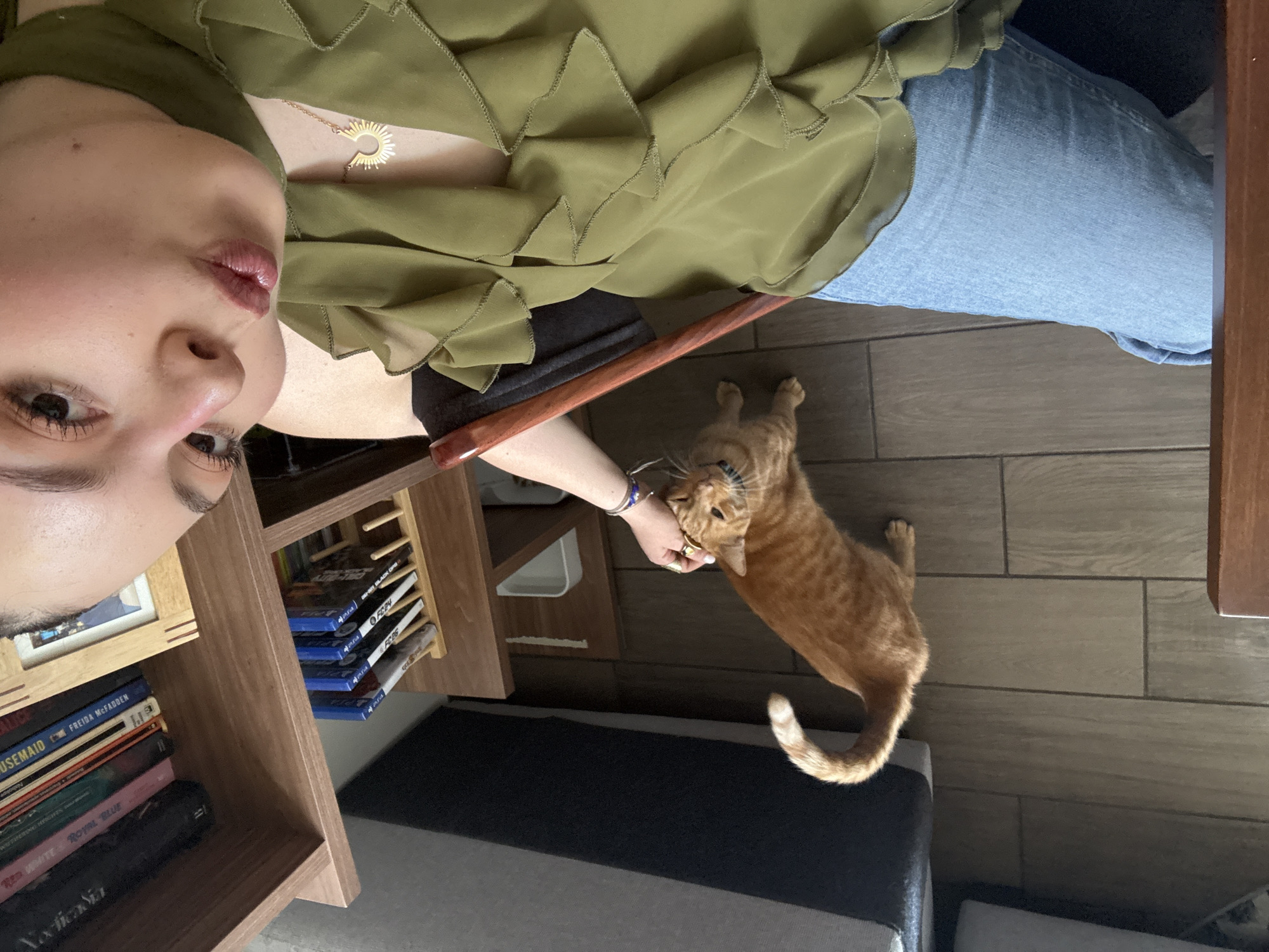 A selfie-style, medium-angle shot depicts a person with light skin, wearing a green ruffled top and light blue jeans, seated in what appears to be an indoor setting. To the left, a wooden bookshelf is partially visible, holding a stack of books and some video game cases. A ginger tabby cat stands on a dark tile floor, interacting with the person's hand. The environment suggests a home interior with neutral-toned walls. There is no information available in this image that would indicate this location to be in Mexico City, Mexico.