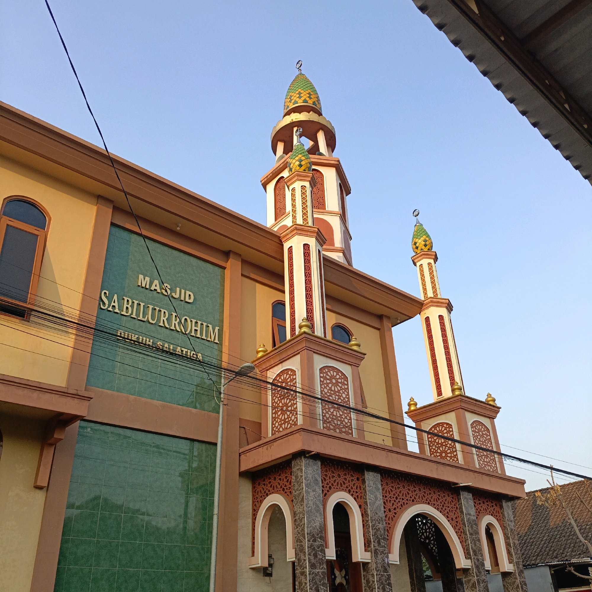 A low-angle, full shot captures the front and side of the Masjid Sabilurrohim in Salatiga, Indonesia, under a clear, pale blue sky. The mosque features a cream-colored facade with brown trim. To the left, a section of the wall is covered in dark green square tiles, with the name "MASJID SABILURROHIM" inscribed in raised, gold lettering. Below this, the text "BUKUN SALATIGA" is also present. A brown-framed, arched window is visible on the left side. The right side of the mosque is dominated by a prominent entrance with arched openings, decorated with intricate geometric patterns in red and white. Flanking the entrance are two square pillars with detailed cutouts. Two towers, or minarets, rise above the main structure. The taller, central tower has a red and white base and tapers upwards to a golden platform supporting a multi-colored, green and yellow patterned dome topped with a crescent moon. A smaller, similar tower is visible to the right. Electrical wires are strung across the scene, and a street lamp is present near the entrance. A portion of a tiled roof of a neighboring building is visible in the upper right corner. The lighting suggests it is late afternoon, with the sun casting a warm glow on the mosque's exterior. No people are visible in the image.