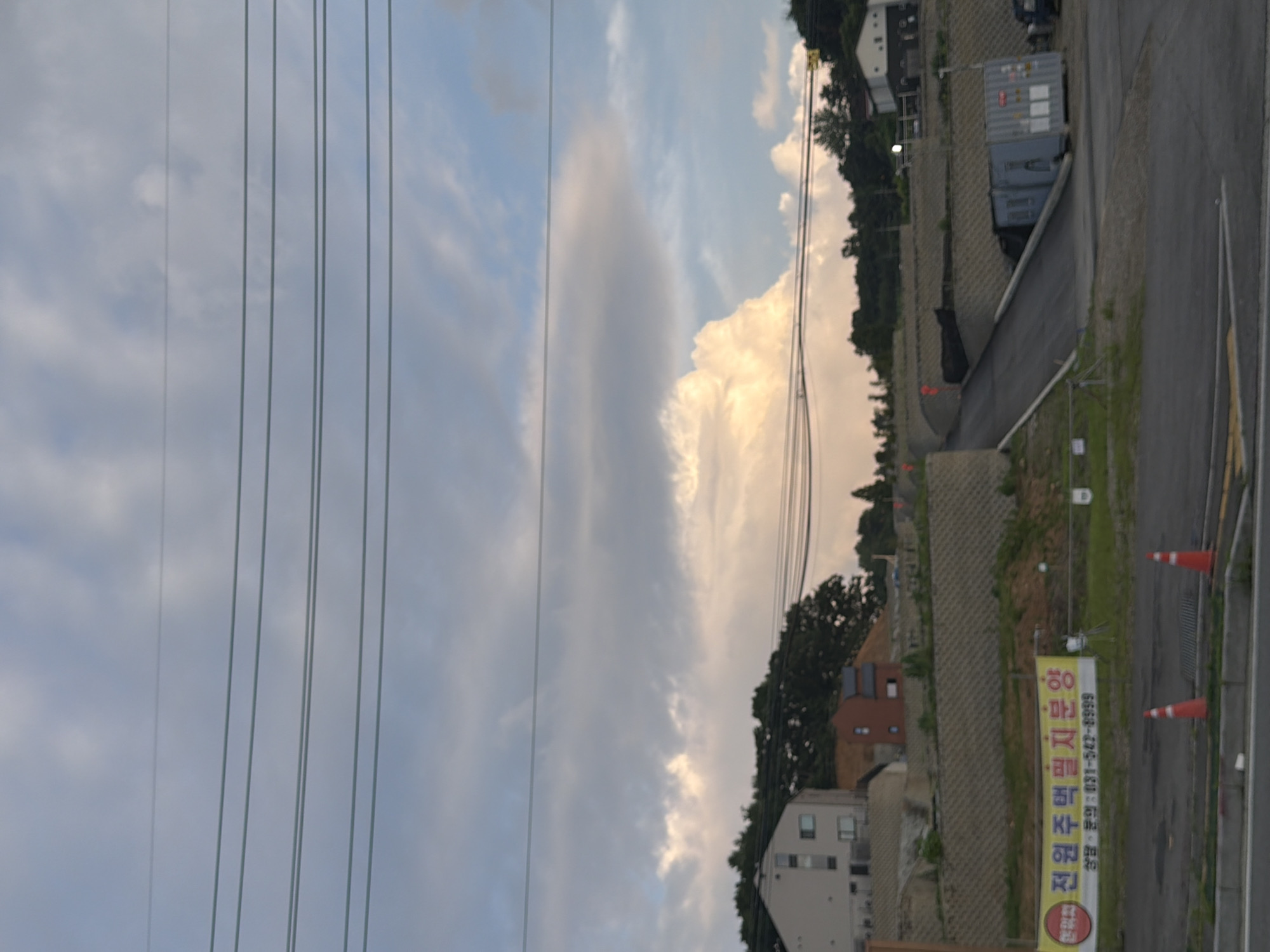 The image depicts a construction site in Pocheon-si, South Korea, under a cloudy sky with visible power lines in the foreground. The landscape is characterized by tiered, terraced earth retaining walls constructed with concrete blocks, supporting a sloped road leading upwards. A building complex with multiple units is situated on the left, and on the right, a large blue shipping container is parked near the road. A yellow banner with Korean text, reading "전원주택부지분양" (which translates to "Rural House Site for Sale") and a phone number, is prominently displayed on the left side of the image. Orange traffic cones are placed on the road. The overall scene suggests ongoing development in a semi-rural or suburban area.