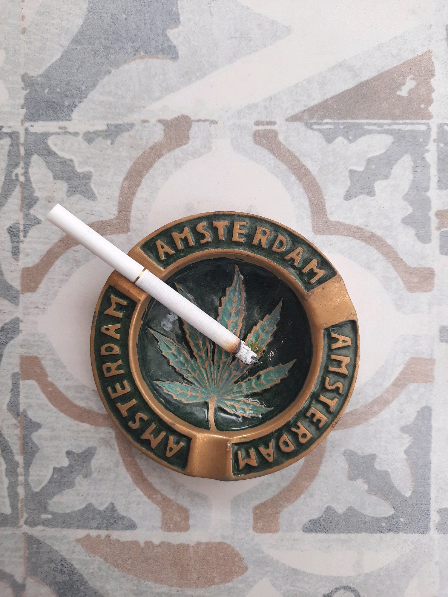 A close-up, overhead view shows a cigarette resting in a round ashtray with a cannabis leaf design. The ashtray is golden-brown with a dark green interior featuring the leaf and the word "AMSTERDAM" embossed around the rim. The cigarette is lit, with ash at its tip. The ashtray is placed on a tiled surface with a patterned design of grey, beige, and blue shapes. There are no people visible in the image. This image depicts a scene that is not tied to any specific location in Novorossiysk, Russia, or any other geographical area.