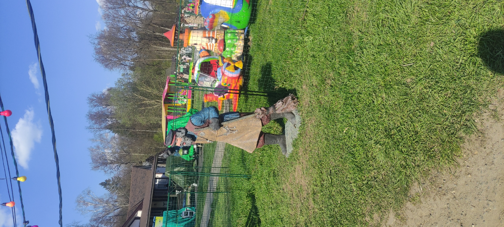 The image depicts an outdoor scene in Grebnevo, Russia. A green metal fence encloses an area with a small amusement park visible in the background, featuring colorful, cartoon-themed rides. In the foreground, there is a decorative statue of a man dressed in old-fashioned clothes. The statue stands in a grassy area, with a dirt path to the right. Trees and a building with a brown roof are visible in the distance. Overhead, power lines are strung with multi-colored light bulbs. The sky is blue with scattered white clouds.
