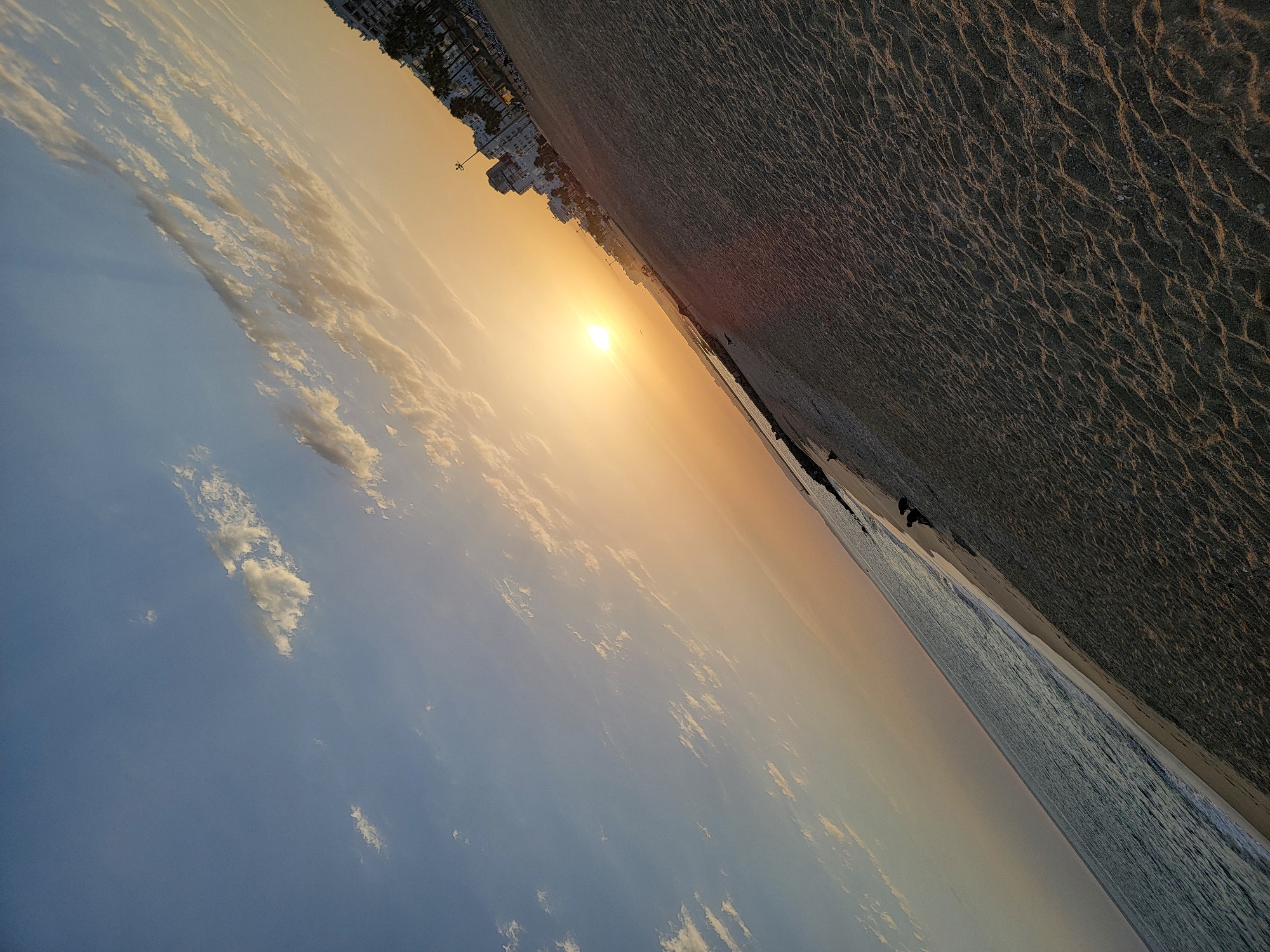 The image captures a coastal scene in Quarteira, Portugal, during a sunset. The sun is low on the horizon, casting a warm orange glow across the sky and reflecting on the calm ocean. The beach, covered in sand with visible ripples, occupies the foreground and midground, stretching towards the right of the frame. Several small birds are scattered on the sand. In the distance, along the coastline, are buildings and palm trees, indicating an urbanized area. A jetty extends into the water on the left side of the image. The overall impression is one of tranquility as day transitions to evening.