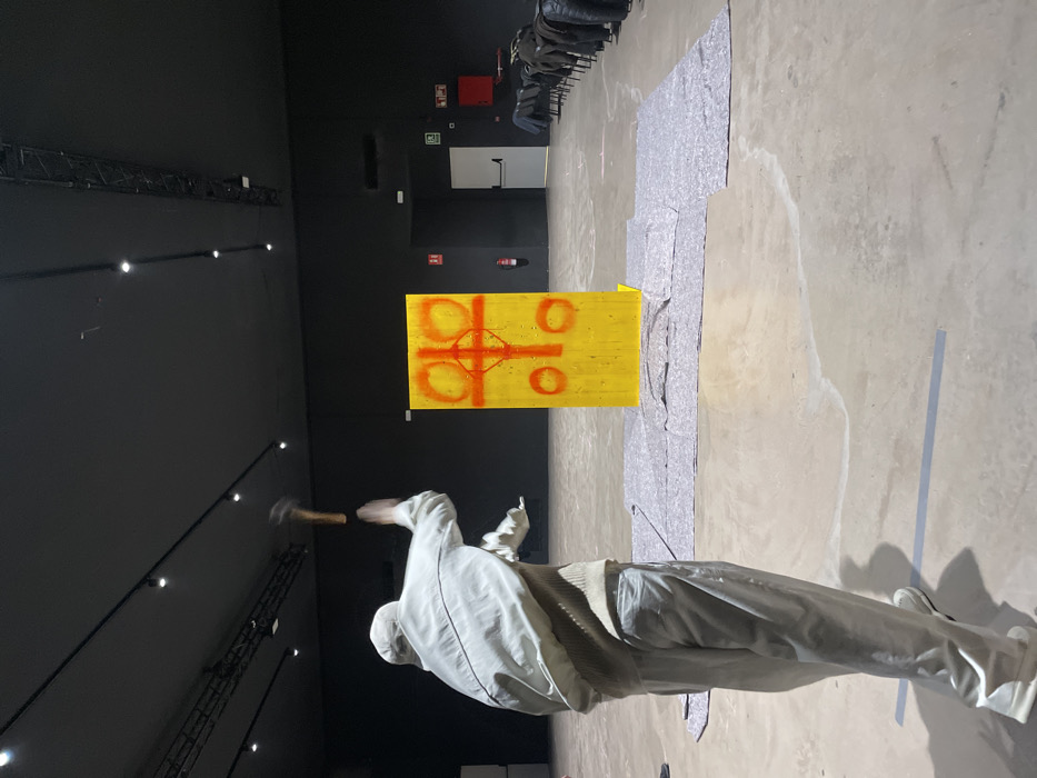 The scene depicts an interior view of a large, dimly lit exhibition space, consistent with gallery areas found at the Fundació Joan Miró in Barcelona, Spain. A person, observed from behind, is captured in motion, actively swinging a hammer with both hands held high above their head, poised for a downward strike. This individual is dressed in light-colored, possibly off-white, clothing consisting of a jacket, sweater, pants, and white shoes. Their target is a freestanding yellow rectangular panel positioned in the mid-ground, marked with abstract red spray paint designs, including a central cross-like figure with two large circular elements on either side and two smaller ones below.
The concrete floor in front of the yellow panel is covered with several grey, patterned fabric sheets. A silver adhesive tape line runs across the foreground floor. The room features a high black ceiling equipped with multiple track lighting fixtures. In the background, along the right wall, a row of dark chairs is visible. Further back, on a dark wall, a white door, a darker doorway, and a red fire extinguisher cabinet with associated white and green safety signage are present.