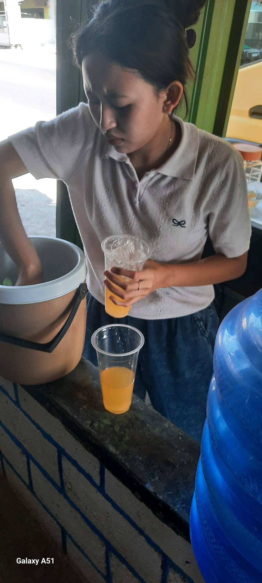In Selopuro, Indonesia, a young woman with her hair pulled back in a bun is focused on preparing drinks. She is wearing a light-colored polo shirt and dark blue pants. Her left hand is submerged in a large, light brown bucket, suggesting she is either mixing or serving. In her right hand, she holds a clear plastic cup filled with a yellow beverage and ice. Another identical cup, also containing the yellow drink, sits on a dark, weathered surface in front of her. To the right of the frame, a large, bright blue water dispenser is partially visible. The background is blurred, but hints of green and yellow suggest an outdoor setting, possibly a market or roadside stall. The overall impression is one of a casual, everyday scene of beverage service.