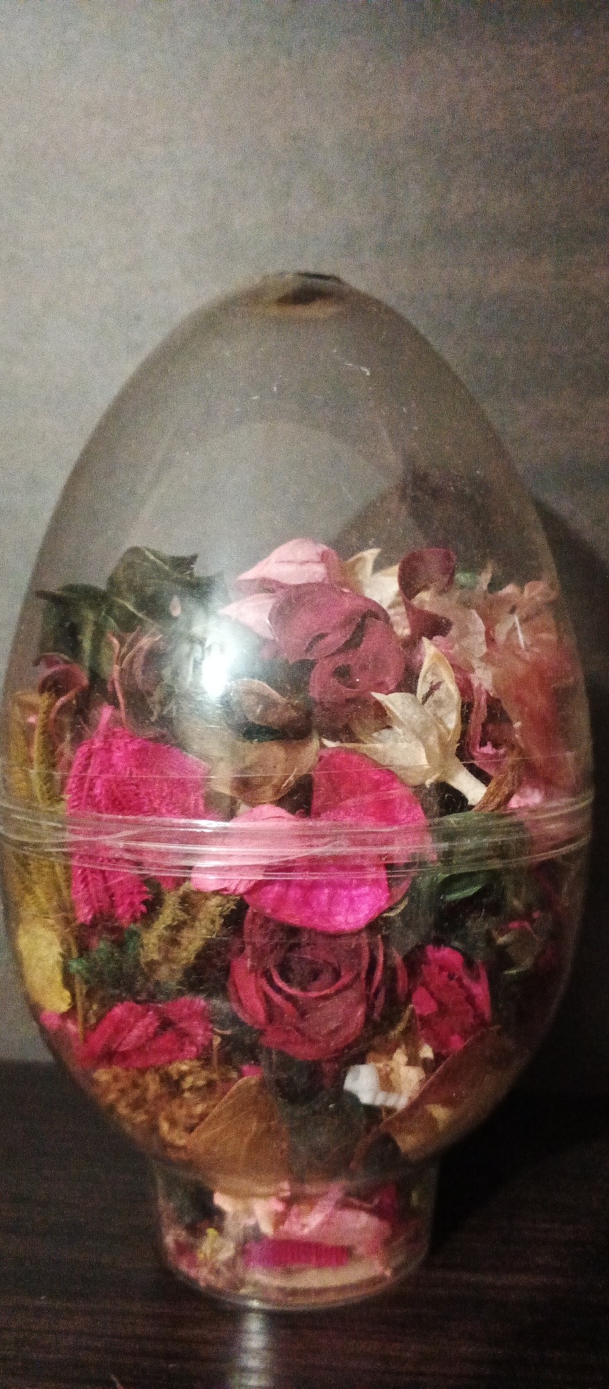 A clear, egg-shaped glass container, filled with potpourri, sits on a dark wooden surface against a gray, textured background. The potpourri consists of dried rose petals in various shades of pink, red, and beige, interspersed with green leaves and other dried floral elements. The container has a slight indentation at the top and a rim around the middle, where the top and bottom halves are joined. The lighting casts a subtle reflection on the glass. The scene does not depict any specific urban environment or location within Berehove, Ukraine.