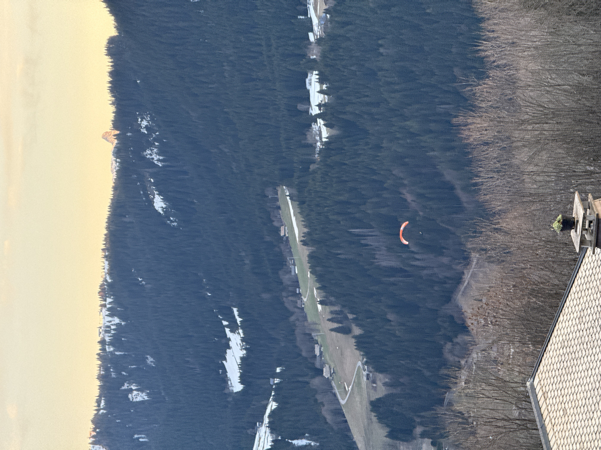 The image shows a view in Morzine, France. In the center of the frame, a red paraglider is in flight above a forest with scattered patches of snow. A narrow, winding path, possibly a road or clearing, cuts through the forest. A building, partially visible in the bottom right, has a light-colored tiled roof. In the background, a mountain range is discernible, with a snow-covered peak on the left. The sky is bright, suggesting either early morning or late afternoon lighting. The scene captures a landscape with elements of nature and human activity.