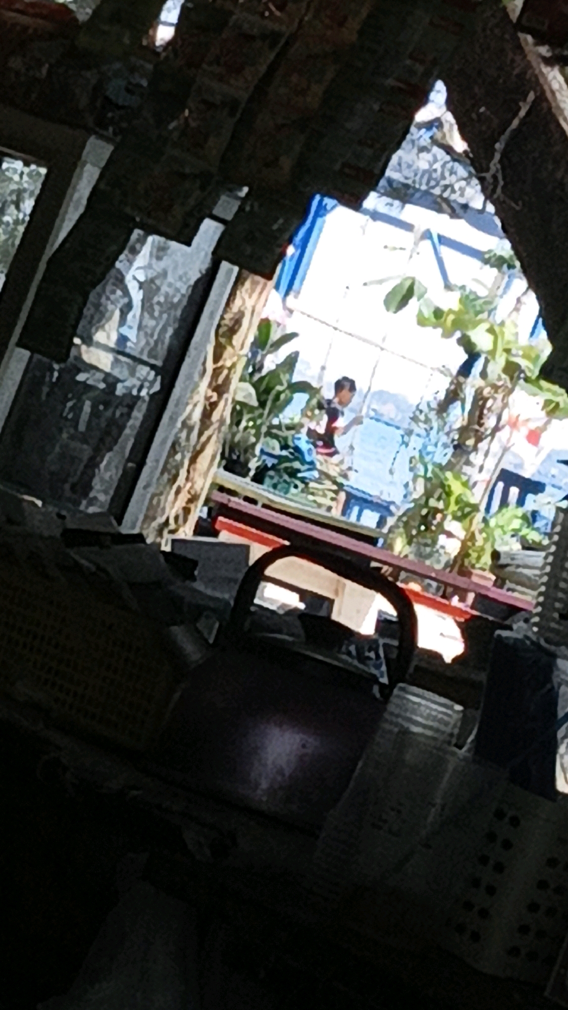 The image depicts an interior view through a damaged or open structure, possibly a shelter or a partially constructed building, offering a glimpse of an outdoor scene. The foreground is dominated by dark, shadowy objects, including what appears to be a woven basket and a metallic, rounded object, possibly a pot or kettle. The midground reveals a person, appearing to be a young male, standing on a platform or elevated area. He is wearing a patterned shirt and is looking towards the water. The background features a body of water, likely the sea, and a distant shoreline with vegetation. The overall impression is of a rustic or informal setting. The location, Cikal, Indonesia, suggests a tropical or coastal environment, which aligns with the presence of the water and vegetation. The damaged structure in the foreground could indicate an area affected by natural elements or ongoing construction.