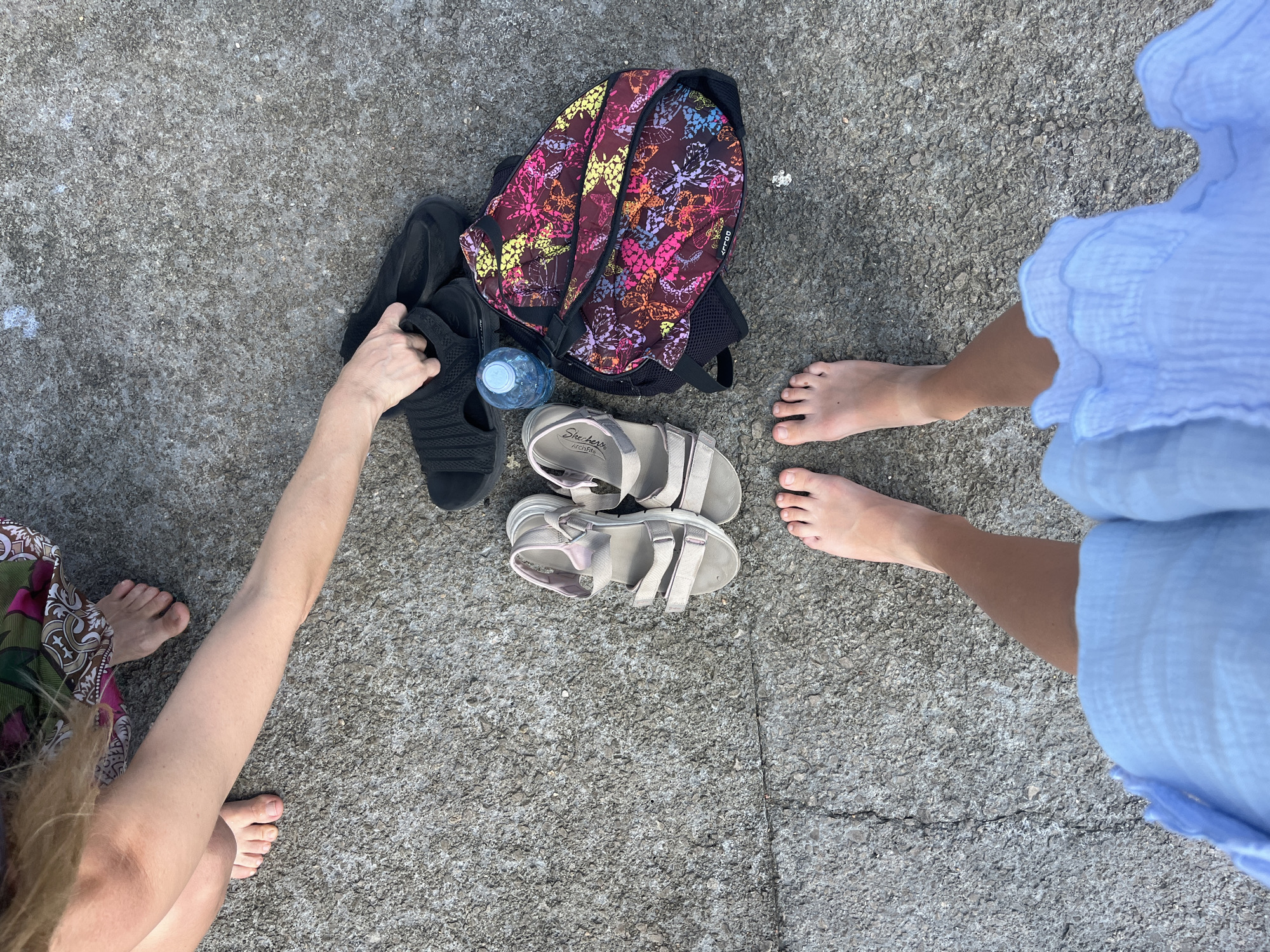 A top-down shot shows two pairs of feet and various items on a grey, textured surface that appears to be a rock or pavement. One person, whose feet are visible on the left, is wearing a patterned skirt. They are reaching for a pair of black slides. Beside the slides is a backpack with a colorful butterfly pattern and a partially full water bottle. A second person, whose feet are on the right, is wearing a light blue ruffled dress. Between the two people are a pair of beige-strapped sandals. The lighting suggests it is daytime, likely sunny. The scene appears to be outdoors, possibly near a beach or waterfront, given the casual attire and the mention of Budva, Montenegro, a coastal town. There are no explicit emotional cues, but the arrangement of items suggests preparation for an outdoor activity or a break from one. The visible text on the sandals reads "Skechers Arch Fit".