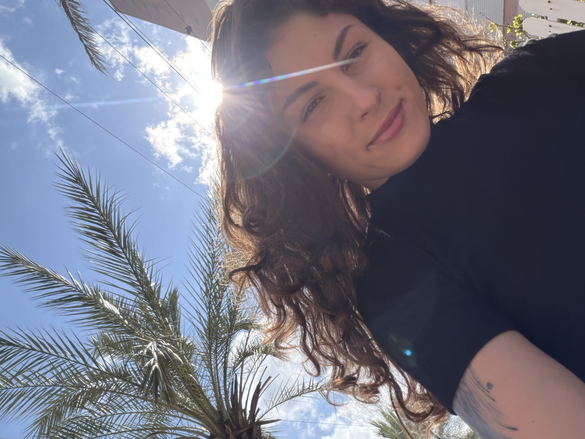A medium shot shows a woman in a black, short-sleeved shirt, angled slightly downward toward the camera with sunlight glaring in the upper left of the frame. Her brown, curly hair is loose around her face. A palm tree appears in the lower left corner. The sky, blue with scattered clouds, is visible in the background. The image appears to have been taken outside in Kiryat Bialik, Israel, judging from the palm trees and building style.