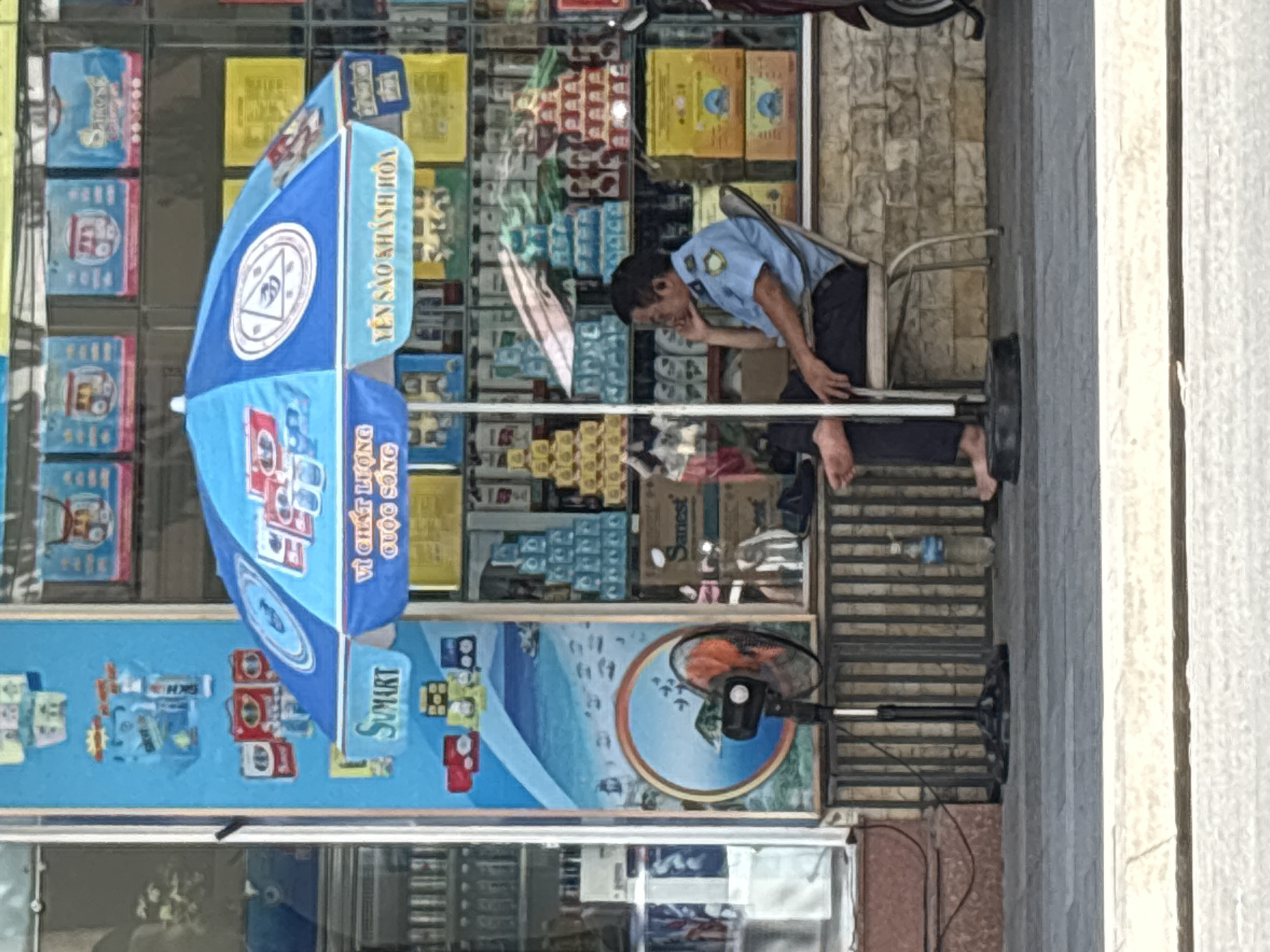 The image depicts a street scene in Nha Trang Ward, Vietnam, with a man in a blue uniform sitting on a chair under a blue umbrella. The umbrella has advertisements with Vietnamese text. The man is leaning his head on his hand, with his bare feet visible. To his left, a black fan with orange blades is positioned on a stand. Behind the man and umbrella, a storefront is visible, displaying rows of stacked products, predominantly in blue, yellow, and red packaging. Some of these packages appear to be drinks or food items. The storefront itself has a textured, stone-like facade. A motorcycle is partially visible to the right of the frame. The ground in front of the storefront is paved with grey tiles, and a white tiled border is visible in the foreground.