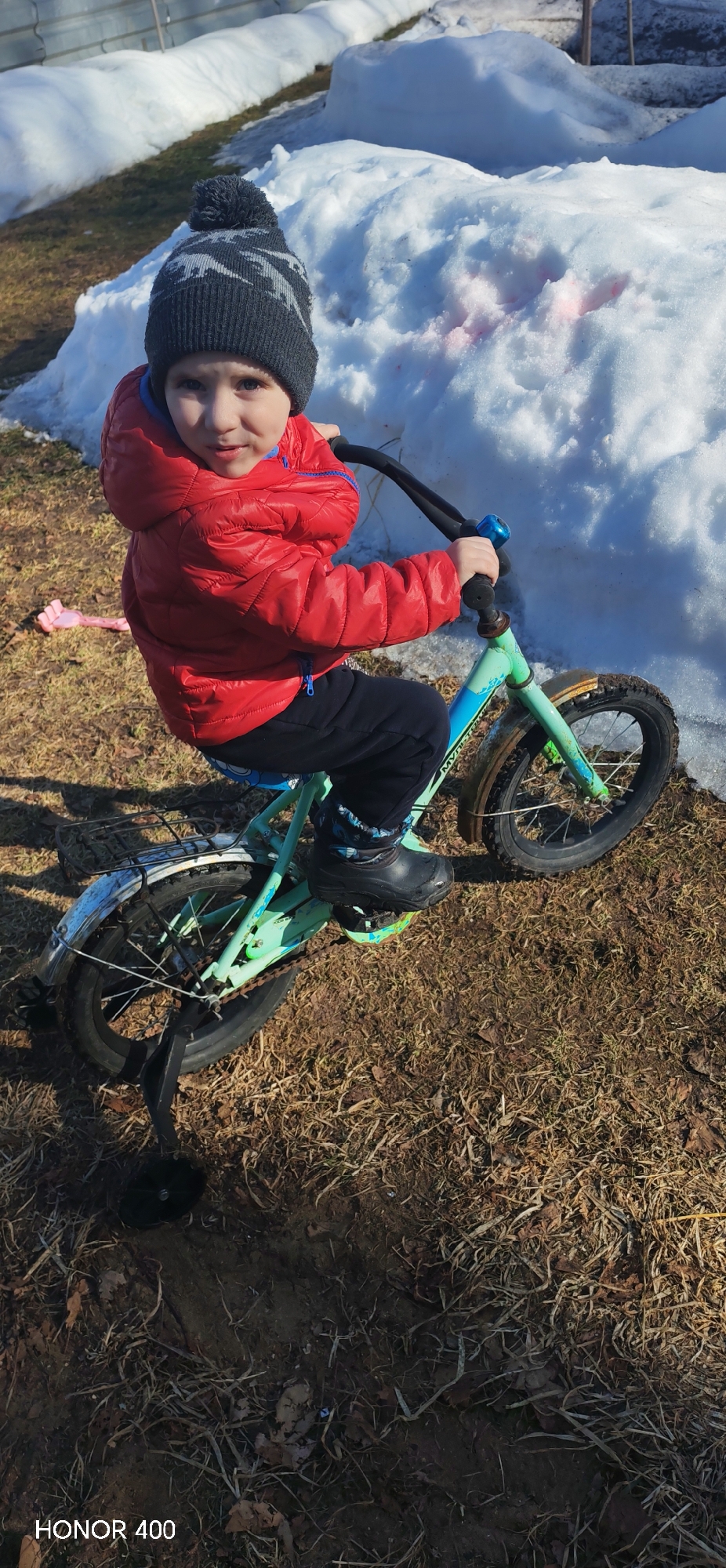 An outdoor scene in Noviy, Russia, depicts a young child seated on a light blue-green bicycle. The child, wearing a dark grey knit hat with a pom-pom and a lighter grey pattern, a red hooded puffer jacket with blue trim, dark blue pants, and black boots, is positioned centrally and looks directly at the camera. The bicycle has black handlebars, black tires, silver mudguards (the front one appearing rusty), and a blue bell attached to the handlebars. A single black training wheel is visible on the left rear of the bicycle. The ground beneath the child and bicycle consists of dry, brown grass, exposed soil, and scattered fallen leaves. To the right and behind the child are irregular, melting patches of snow, one of which exhibits pink discoloration. A pink plastic object is visible on the ground to the left of the child. In the distant background, beyond the snow, a fence or structural element is partially visible under bright, sunny conditions.