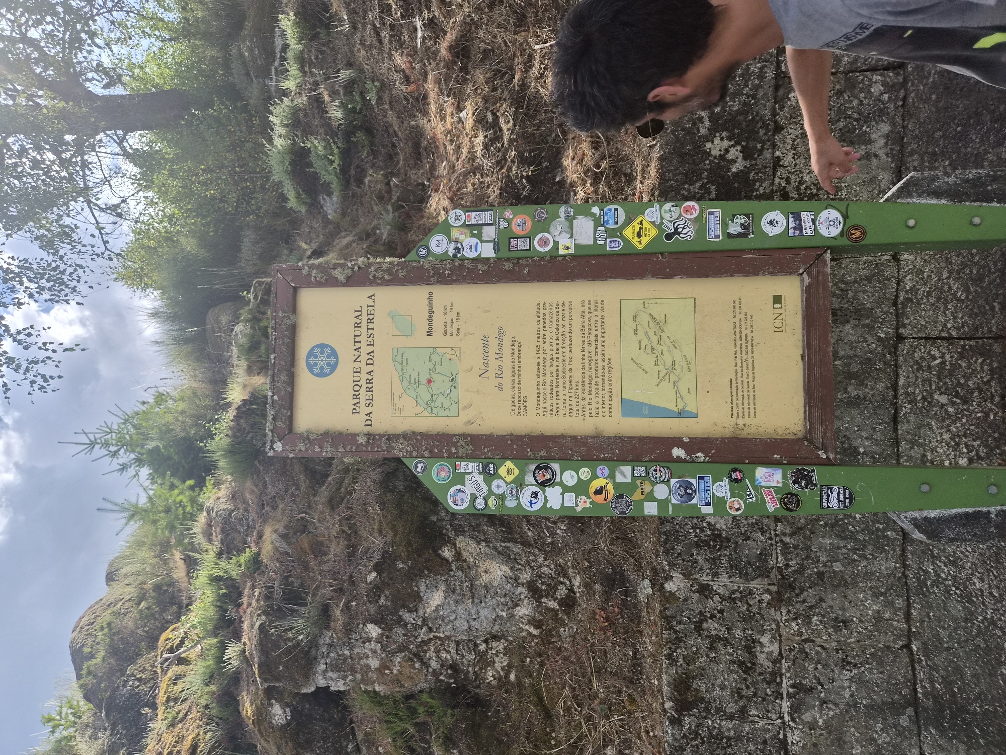 The image depicts an outdoor scene featuring a directional signpost adorned with numerous stickers, situated against a backdrop of a rocky, vegetated hillside. In the foreground, a man in a gray t-shirt, with sunglasses on, is seen from the side, pointing at the signpost. The signpost itself is green and rectangular, with a large informational panel at the top. The panel contains text and a map, with "PARQUE NATURAL DA SERRA DA ESTRELA" prominent at the top. Text below indicates locations such as "Mondegusinho" and "Rio Mondego," suggesting a connection to the Serra da Estrela Natural Park, which is located in the municipalities of Gouveia, Covilhã, and Manteigas in Portugal. The man's gesture implies he is either explaining something on the sign or drawing attention to it. The environment is natural, with dry vegetation and rocks, under a bright sky with some clouds.