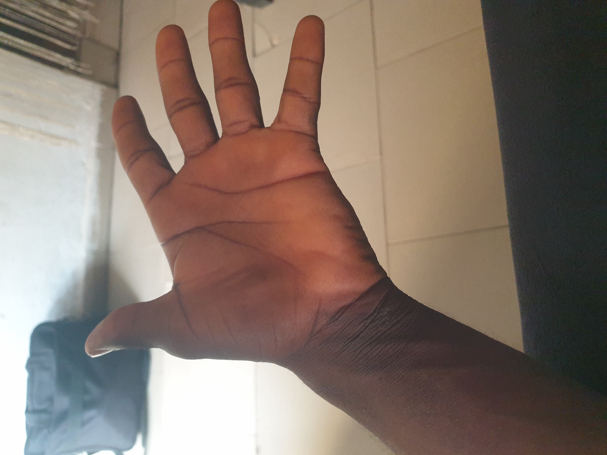 The image shows a close-up view of an open right hand, palm facing upwards. The hand exhibits dark skin tone with visible palm lines and creases. The fingers are spread apart, with the thumb slightly extended. The background is blurred, but appears to be an indoor setting with tiled flooring and a wall. A dark bag is visible in the upper left corner of the frame, and a portion of a dark surface is in the lower foreground.