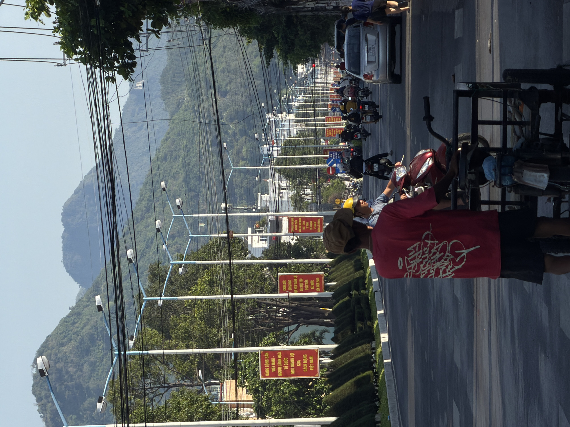 A street scene in Bac Nha Trang Ward, Vietnam, features a road lined with trees and lampposts. Mountains form the backdrop. Several people are visible, some riding motorcycles and scooters. One person in the foreground, wearing a red t-shirt and black shorts, stands with their back to the camera, next to a three-wheeled vehicle. Other individuals, including one wearing a yellow helmet, are also on scooters. A silver car is parked on the right side of the road. Red and white signs with Vietnamese text are posted along the roadside. The overall atmosphere suggests a bustling urban environment under a clear sky.