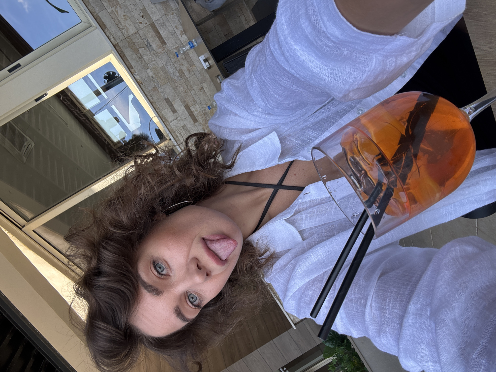 This selfie captures a woman with voluminous, curly brown hair and bright blue eyes playfully sticking out her tongue. She is looking directly at the camera, conveying a cheerful and relaxed mood. She is wearing a lightweight, white linen-style button-up shirt over a black strappy top.
In her right hand, she holds a large, stemmed glass filled with a vibrant orange beverage, likely an Aperol Spritz, complete with ice cubes and two black straws. The glass is positioned prominently in the foreground.
The setting appears to be an outdoor or semi-outdoor area in Prijevor, Montenegro, characterized by bright daylight. In the background, large glass doors or windows reflect a clear blue sky with some scattered clouds, as well as parts of an interior structure. The ground or patio is covered with light-colored, rectangular stone tiles. A table is partially visible with a couple of "Knjaz Milos" branded plastic water bottles on it. Some green foliage is visible at the very bottom right, suggesting nearby plants. A small, faint white sign with "NO..." text is reflected in the glass door.