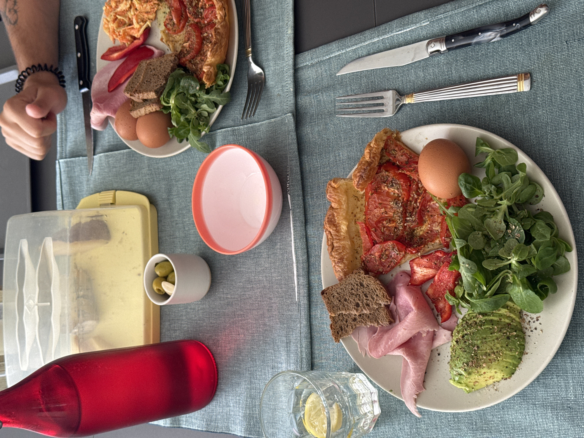 This is a close-up, eye-level shot of a breakfast spread laid out on a blue textured placemat, possibly on a table in Vienne, France. The meal features two plates of food, a bottle of red liquid, a glass of water with lemon, a small bowl of olives, and a pink-rimmed bowl.

The plate in the foreground contains sliced avocado seasoned with pepper, a pile of leafy greens, a slice of torte with tomatoes and herbs, two pieces of dark bread, ham, and a boiled egg. The plate in the background, partially visible, appears to have similar items including ham, bread, two boiled eggs, some shredded carrots, and red pepper slices.

A hand with a black coiled bracelet is visible in the upper right corner, suggesting someone is about to partake in the meal. Utensils, including forks and knives, are placed on the placemat. The lighting is natural, creating soft shadows. The overall scene conveys a leisurely breakfast setting.