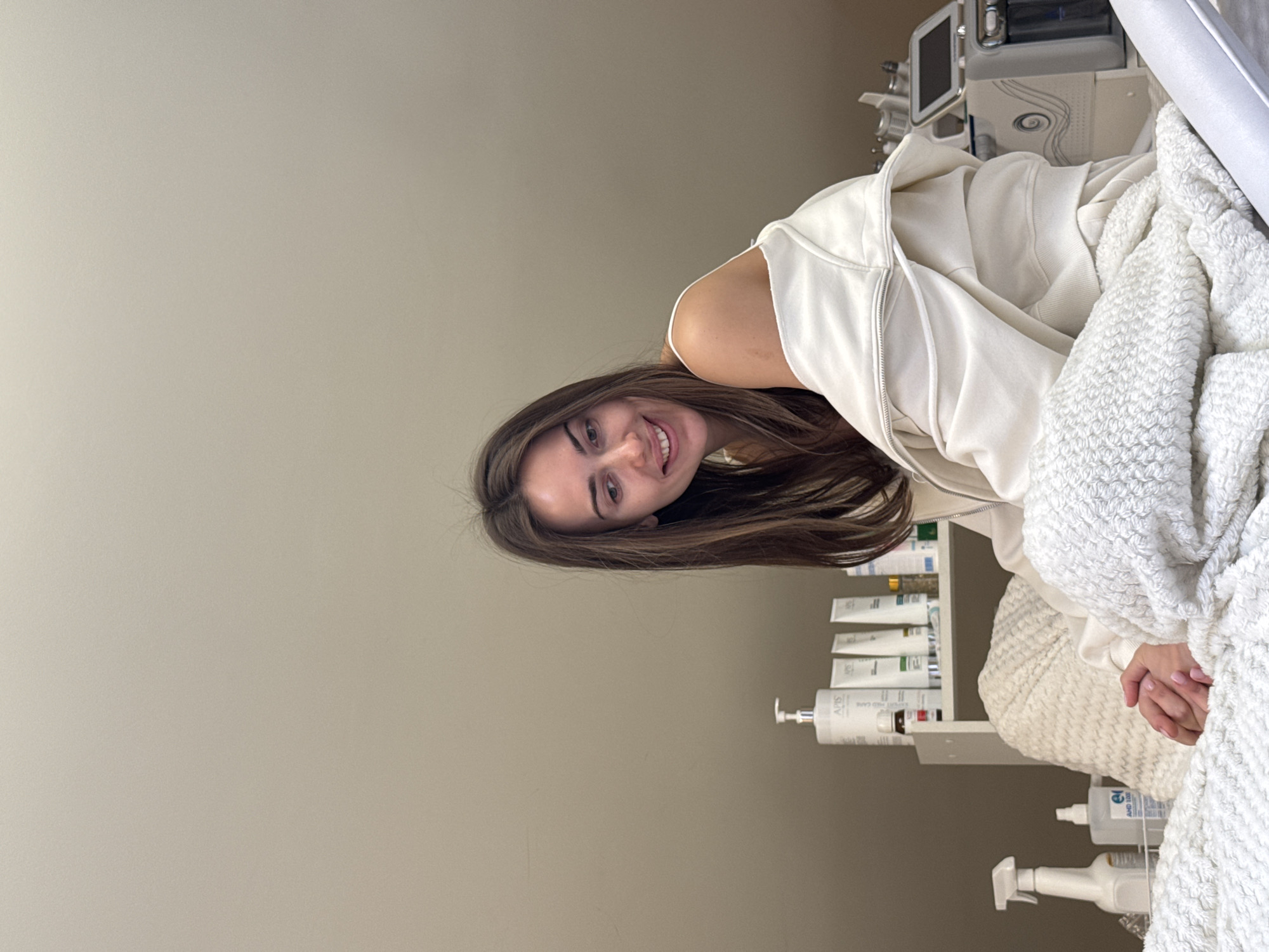 A young woman with long brown hair is lying on a treatment bed in what appears to be a spa or skincare clinic. She is smiling at the camera. She is wearing a white hoodie and is covered with a white, textured blanket. A shelf with various skincare products is visible behind her. Medical equipment is visible in the background. The location is Unknown, Unknown.