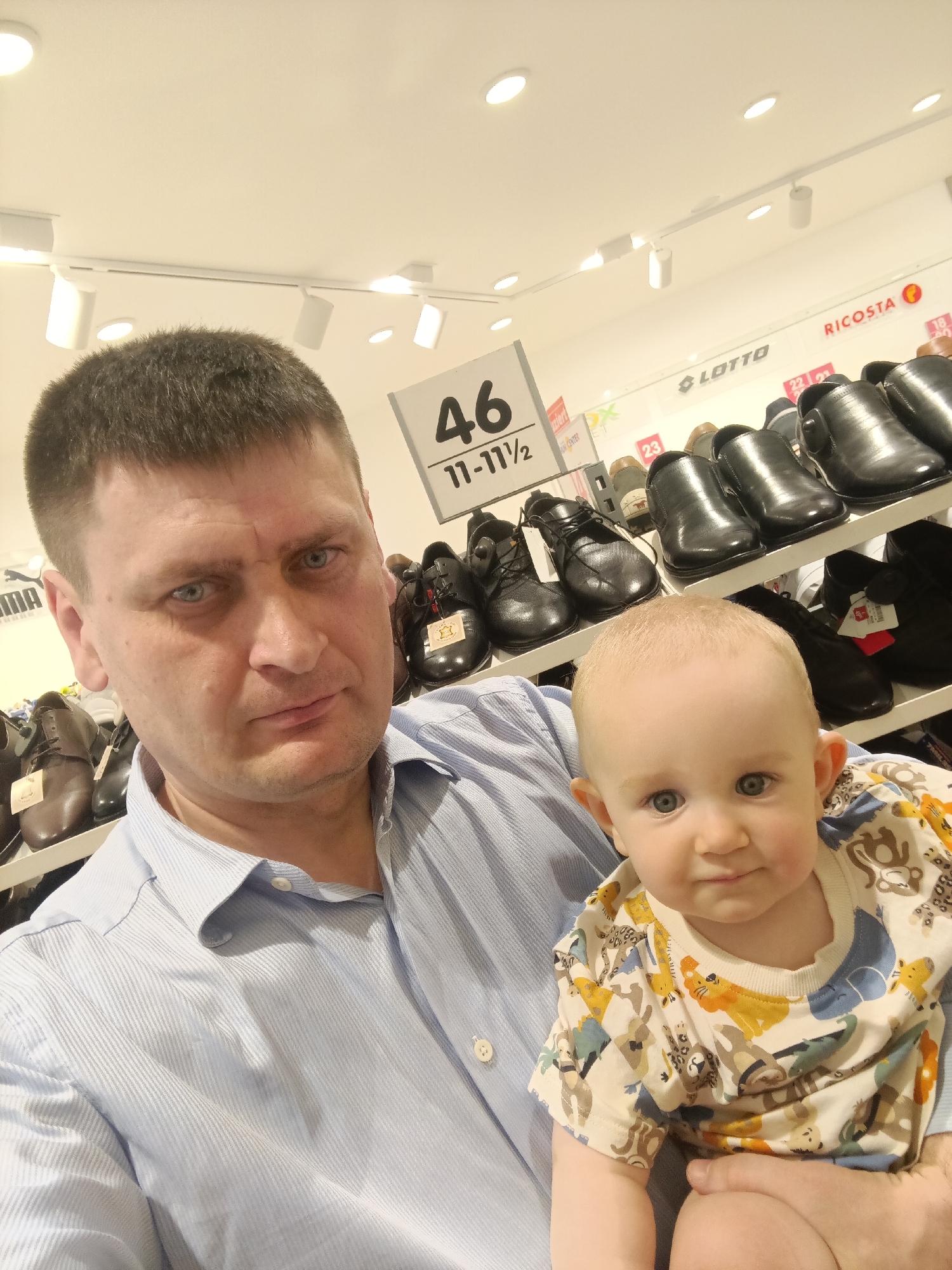A man and a baby are in a shoe store. The man is in the foreground, holding the baby. He is wearing a blue striped shirt. The baby is wearing a white t-shirt with animal prints. Behind them, shelves of shoes are visible. A sign in the background indicates "46" and "11-11½". The store appears to be well-lit. There is no specific indication of the store's location in Nuremberg, Germany.