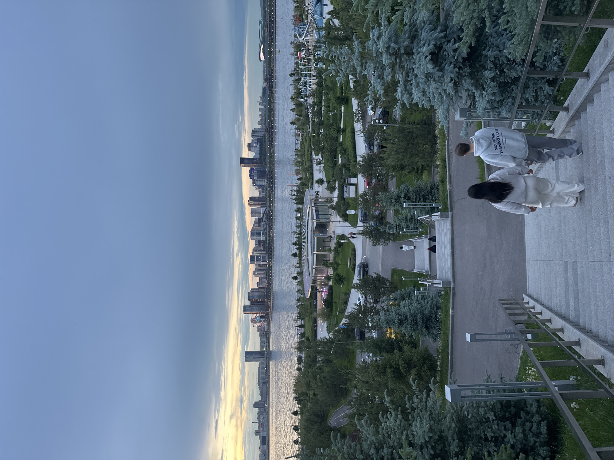 The image shows an elevated view of Kazan, Russia, oriented sideways. A cityscape lines a body of water to the left, under a gradient sky transitioning from blue to sunset hues. Adjacent to the water is a park with pathways, trees, and architectural features. In the foreground, two people are walking down steps. One person is wearing a light-colored jogging suit, and the other is wearing a gray hooded sweatshirt labeled "MOUNTAIN TRAINING CLUB". The steps are bordered by metal railings and vegetation.