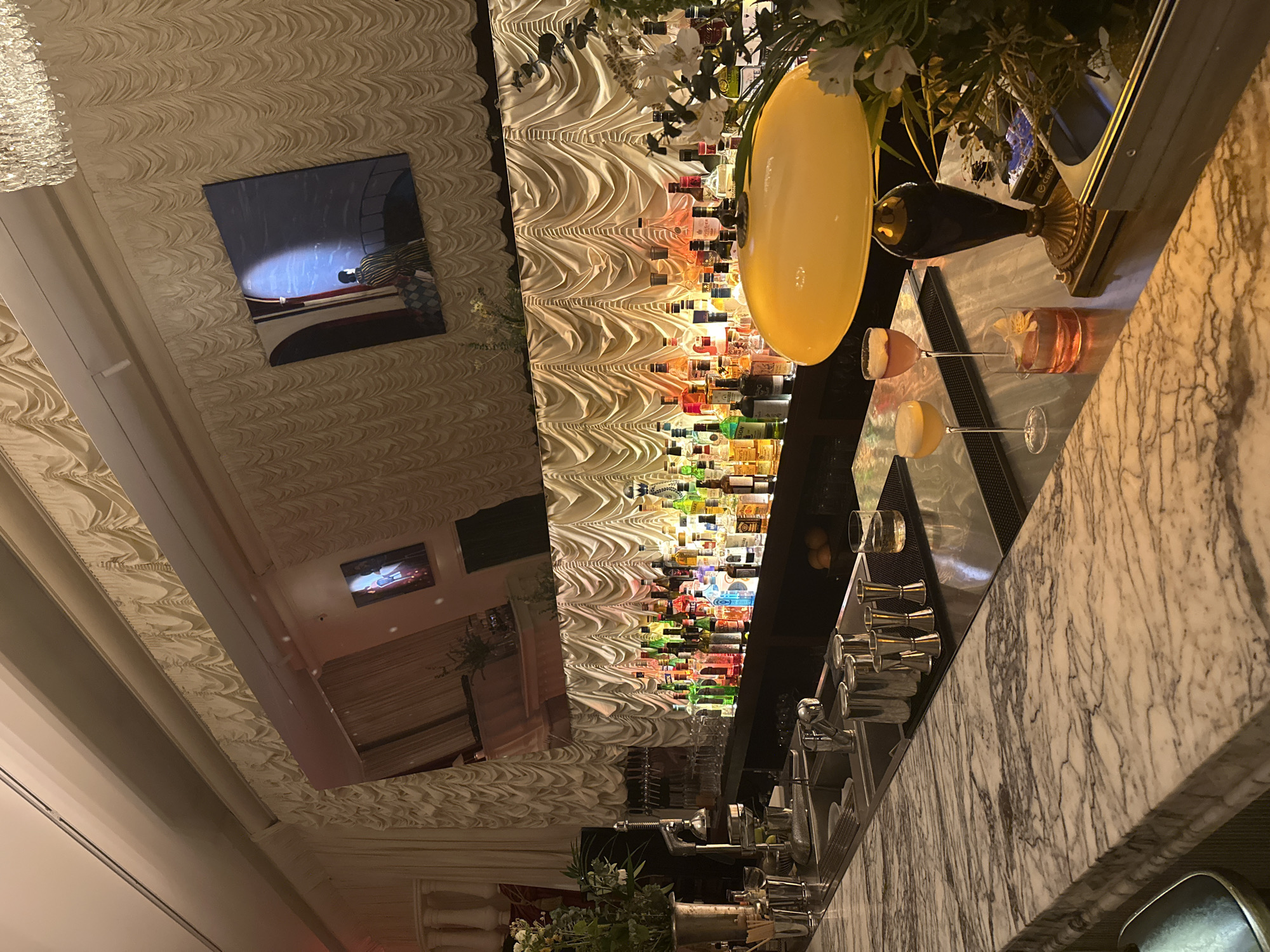 The image shows a dimly lit, opulent bar interior. The bar itself is a long marble counter with a sink and various bar tools, including jiggers and shakers, visible. Behind the counter, a vast array of liquor bottles are neatly arranged on shelves, illuminated from behind by a textured, draped fabric wall in a cream color. A framed artwork depicting a person in a striped coat standing on a bridge is hung on the wall above the bar. On the marble counter in the foreground, there are three cocktails and a vintage-style yellow lamp. The background is further obscured by the draped fabric and a large chandelier, suggesting an elegant establishment. The overall ambiance is sophisticated and upscale.