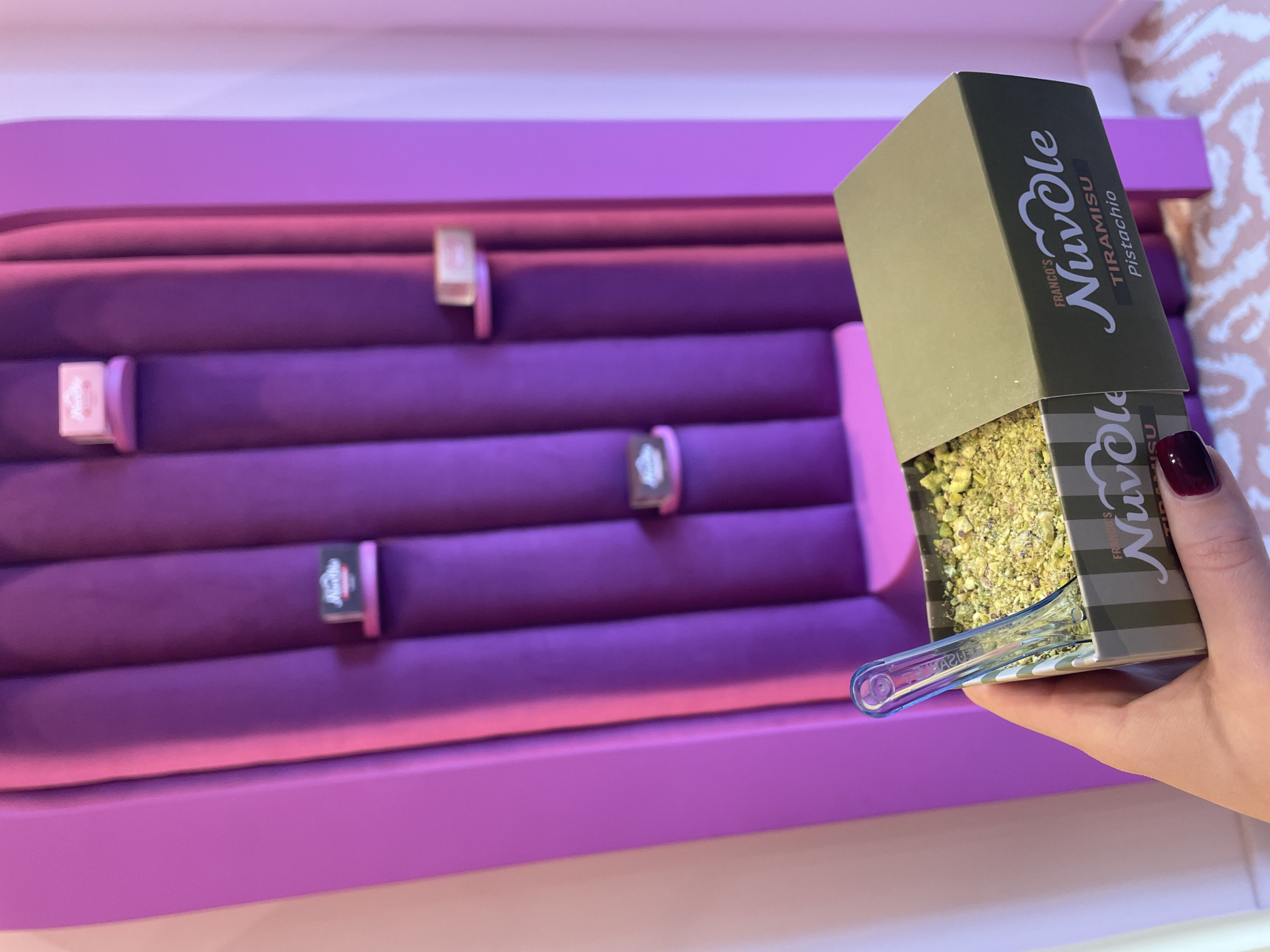 The scene features a partially filled green and black box, labeled "FRANCO'S NuvOle TIRAMISU Pistachio", being held by a person with visible dark burgundy nail polish. The box contains what appears to be pistachio filling, with a small blue utensil partially submerged within it. The background consists of a purple display case with multiple vertical rows, each accented with small, rectangular markers or labels of similar color. In the top right corner of the image, a patterned interior lining is visible within the box. The location is unidentified, and there's no information regarding Sofia, Bulgaria.