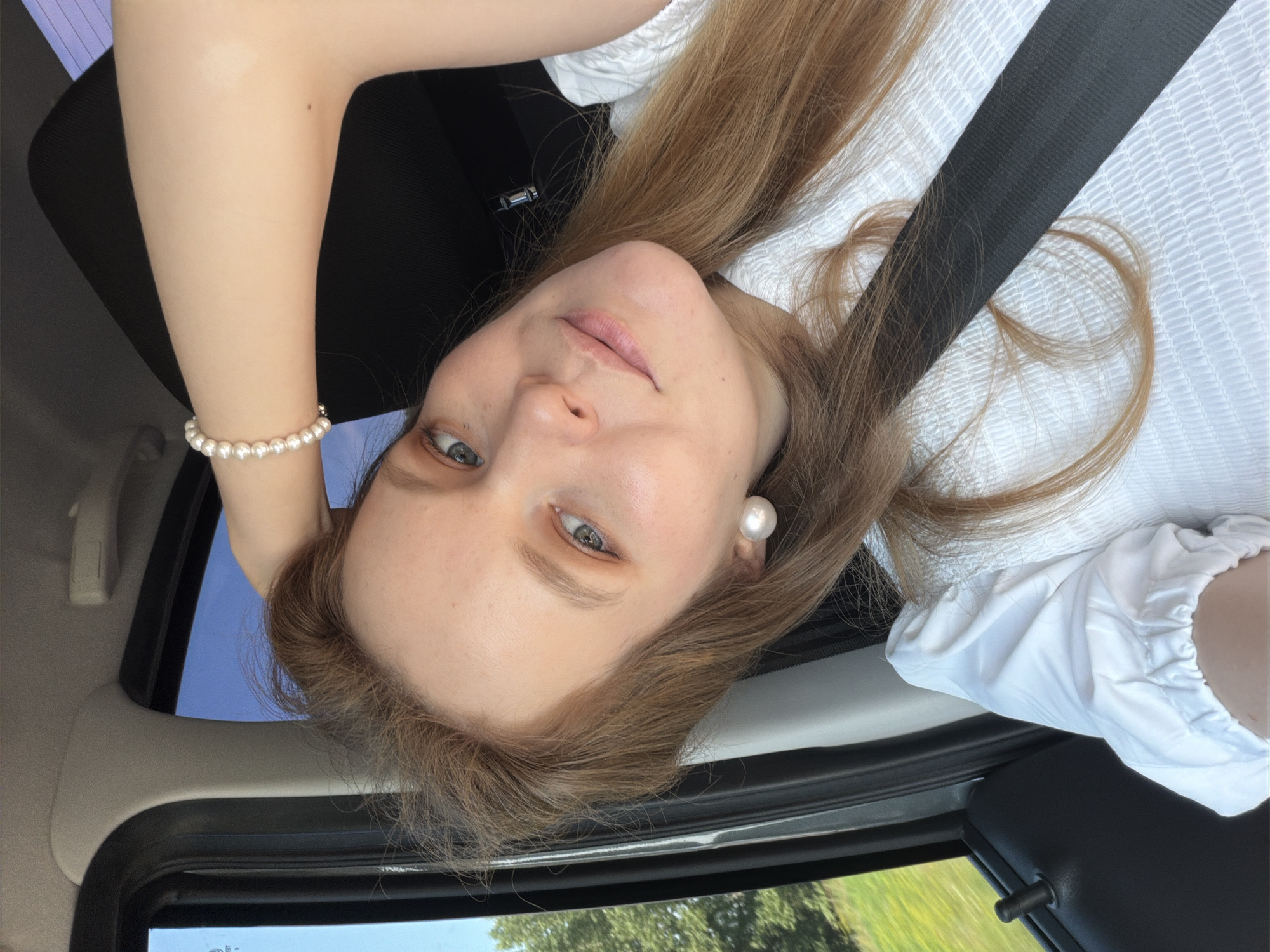 A medium shot captures a woman with long, light brown hair sitting in a car, her right arm raised and hand resting on her head. She is wearing a white, textured shirt and a pearl bracelet. Large pearl stud earrings are visible. Her gaze is directed towards the camera, and her lips are slightly parted. A seatbelt is fastened across her chest. Outside the car window, green foliage and a blue sky are visible, suggesting a daytime setting in a rural or suburban area. The woman's hair is a prominent feature, falling around her shoulders and partially obscuring her right arm. The image appears to be a selfie, taken from a slightly high angle. The location is identified as Tikhonovo, Russia.