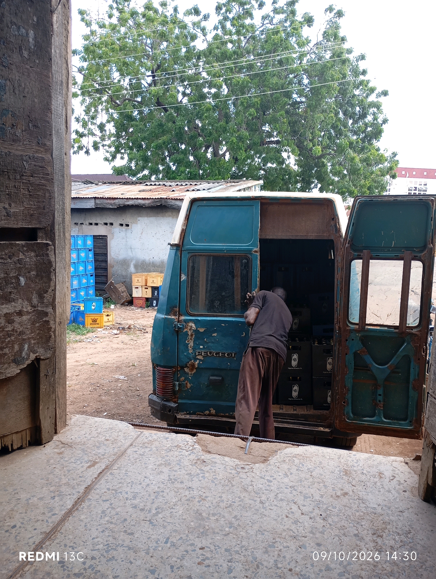 The image depicts an outdoor scene in Shendam, Nigeria, featuring a teal-colored Peugeot van with its rear doors open. A person, wearing a dark t-shirt and brown pants, is seen bending into the back of the van, appearing to be engaged in loading or unloading items. Stacks of blue and yellow crates, some labeled with "NB" and "Guinness," are visible in the background, along with other miscellaneous crates. The environment appears to be a dusty, unpaved area with corrugated metal roofing on a building in the mid-ground. Trees with lush green foliage are present in the background, and power lines are strung across the top of the frame. The foreground consists of a concrete surface with a metal strip running through it. The image is dated 09/10/2026 at 14:30 and branded "REDMI 13C".