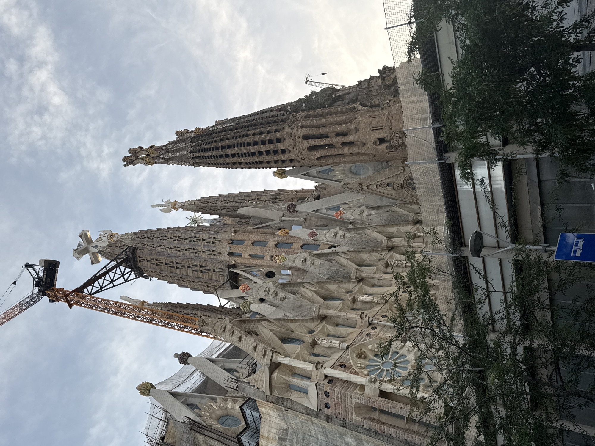 This is a photograph of the Sagrada Familia, a Roman Catholic basilica located in Barcelona, Spain. The image is taken from a low angle, looking up at the spires of the basilica. A large construction crane is visible to the left of the basilica, indicating that construction is ongoing. The sky is cloudy, and the basilica is partially in shadow. Trees are in the foreground, obscuring some of the lower portion of the building. The basilica's distinctive architecture, with its intricate carvings and soaring spires, is clearly visible.