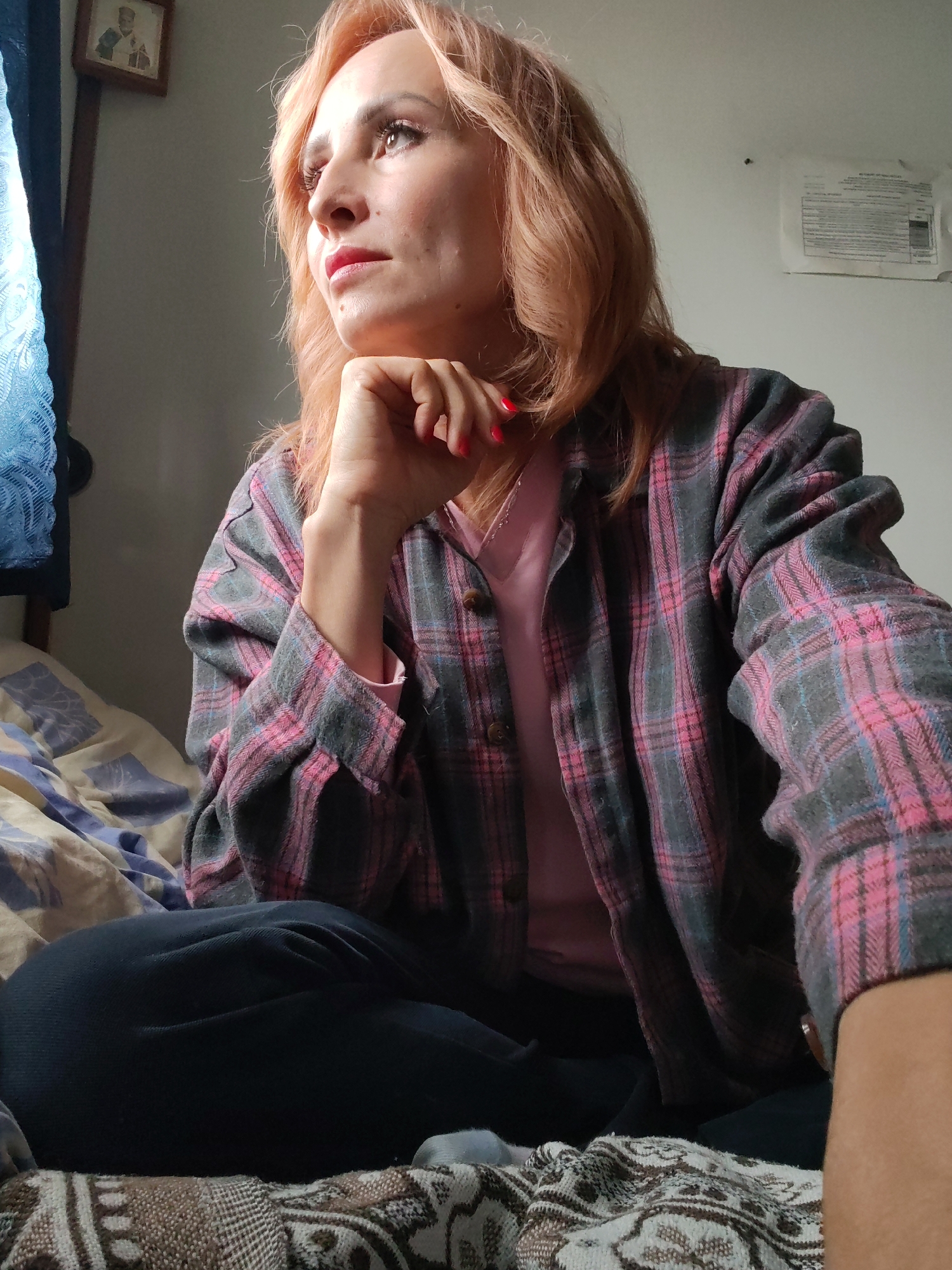 A woman with reddish-blonde hair sits indoors, with her right hand resting on her chin and her gaze directed to the left. She is wearing a pink t-shirt under a grey and pink plaid shirt. She is seated on a bed covered with patterned bedding. To the left of the frame, a blue curtain is visible. In the background, a small framed picture is hung on the wall. The scene appears to be in a domestic setting within the city of Bol'shie Mkhi, Russia.