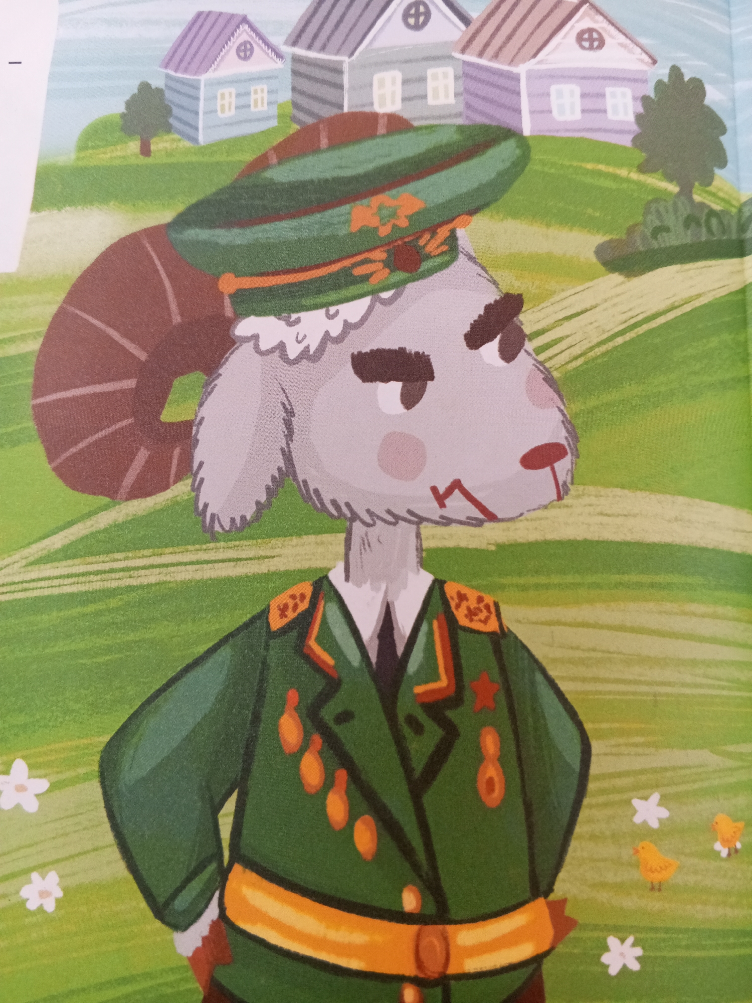 A stylized, cartoonish illustration depicts a ram wearing a military uniform and cap. The ram has light gray fur, large brown curved horns, and is facing to the right of the frame. It is dressed in a dark green military jacket adorned with medals and epaulets, and a green cap with an orange band and emblem. Behind the ram, the background shows a rolling green landscape with several houses in the distance, each with a purple or gray roof. Two small yellow chicks are visible in the lower right corner of the image, and two white stylized flowers are scattered in the foreground. The overall impression is of a children's book illustration, likely from Russia given the context of the city of Chistopol, although the image itself contains no specific markers indicating a city location.