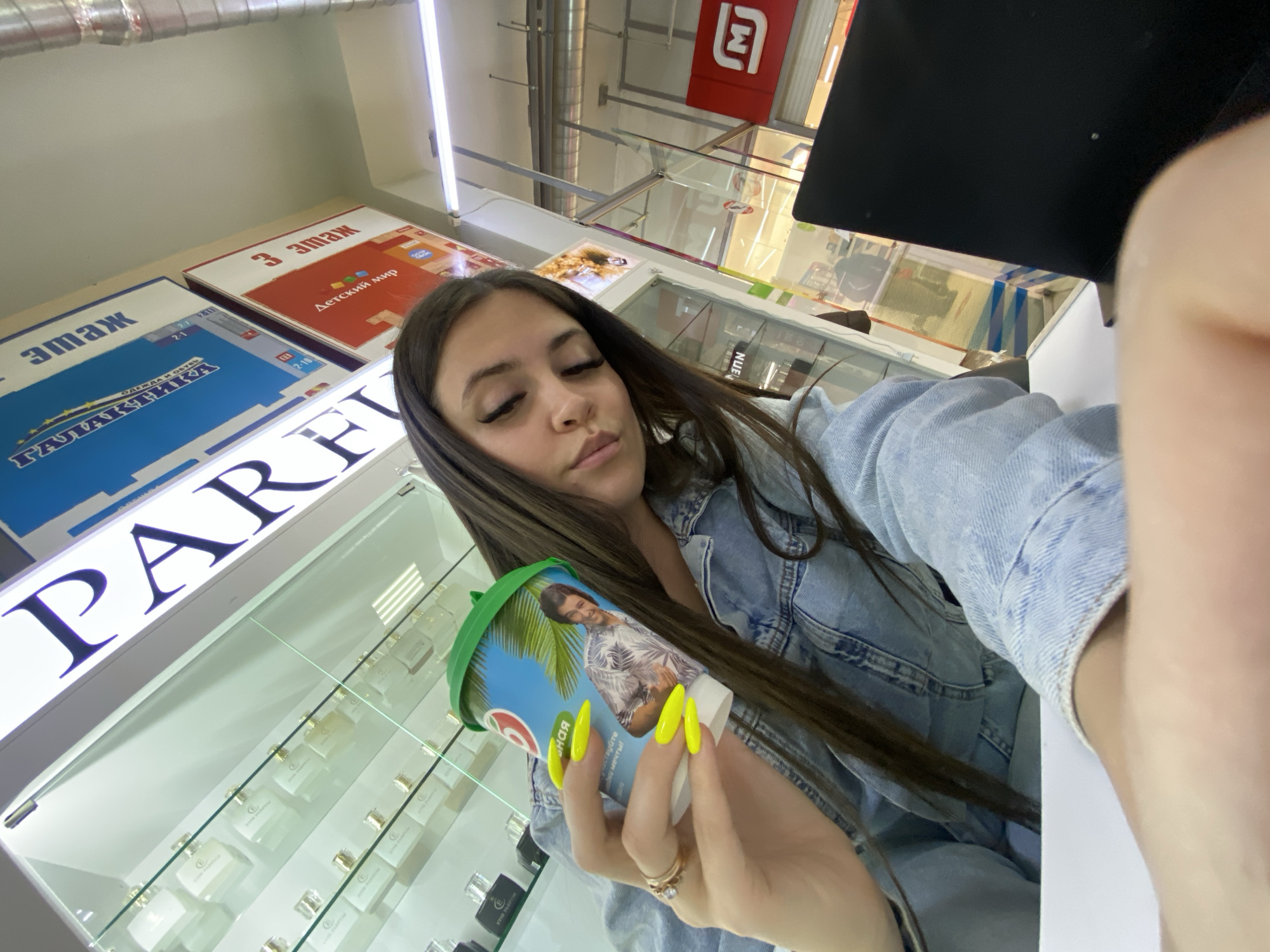 The image appears to be a selfie taken by a woman in what seems to be a commercial establishment in the city of Chaikovsky, Russia. The woman has long dark hair and is wearing a denim jacket. She is holding a cup with an image of a man on it. Her nails are painted a bright yellow.
In the background, there are display cases filled with products, possibly perfumes or cosmetics, judging by the words "PARFI" visible on a sign. The signage in the background indicates the presence of retail areas such as clothing, footwear, and children's goods ("Детский мир" - Children's World). There's also a red logo visible in the background, and a metallic structure, likely part of the building's framework. The store seems to be located on the 3rd floor, according to the signage.