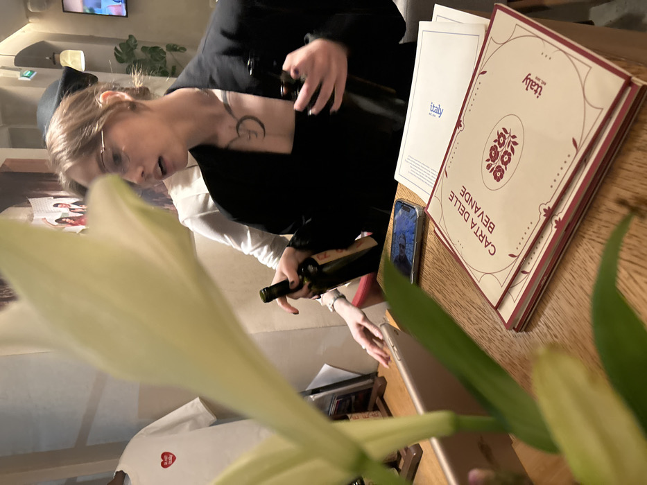 The image captures an indoor dining scene, likely in a restaurant or a home setting, characterized by warm, artificial lighting. The perspective is slightly low, looking across a wooden table with a prominent, out-of-focus white flower (possibly a lily) and green leaves in the immediate foreground on the left.
The central focus is a person, likely a woman, with light hair and wearing glasses. She is dressed in a dark-colored top or jacket over a lighter, long-sleeved shirt. A tattoo, resembling a crescent moon or a stylized letter, is visible on her neck/shoulder area. She is holding two dark-colored bottles, which appear to be wine bottles, suggesting she is either pouring, presenting, or selecting them. Her gaze is directed towards the bottles.
On the wooden table to the right of the person, there are several items. A stack of cream-colored menus is prominently displayed, featuring a decorative floral design in red. The title "CARTA DELLE BEVANDE" (Italian for "Beverage Menu") is printed vertically on these menus, along with the word "italy" and a small logo. Another, simpler white menu with "italy" printed in blue is partially visible beneath the stacked menus.
Also on the table, a smartphone is lying flat, its screen illuminated and displaying the face of a man. Another electronic device, possibly a tablet or a second smartphone, with a lighter-colored casing, is partially visible near the bottom right edge of the frame.
In the background, another person is partially visible, wearing a white long-sleeved shirt with a red heart-shaped logo on it. Further in the background, a television screen is visible on a wall, along with a light fixture and what appears to be another green plant. The overall atmosphere suggests a relaxed gathering or a meal being enjoyed.