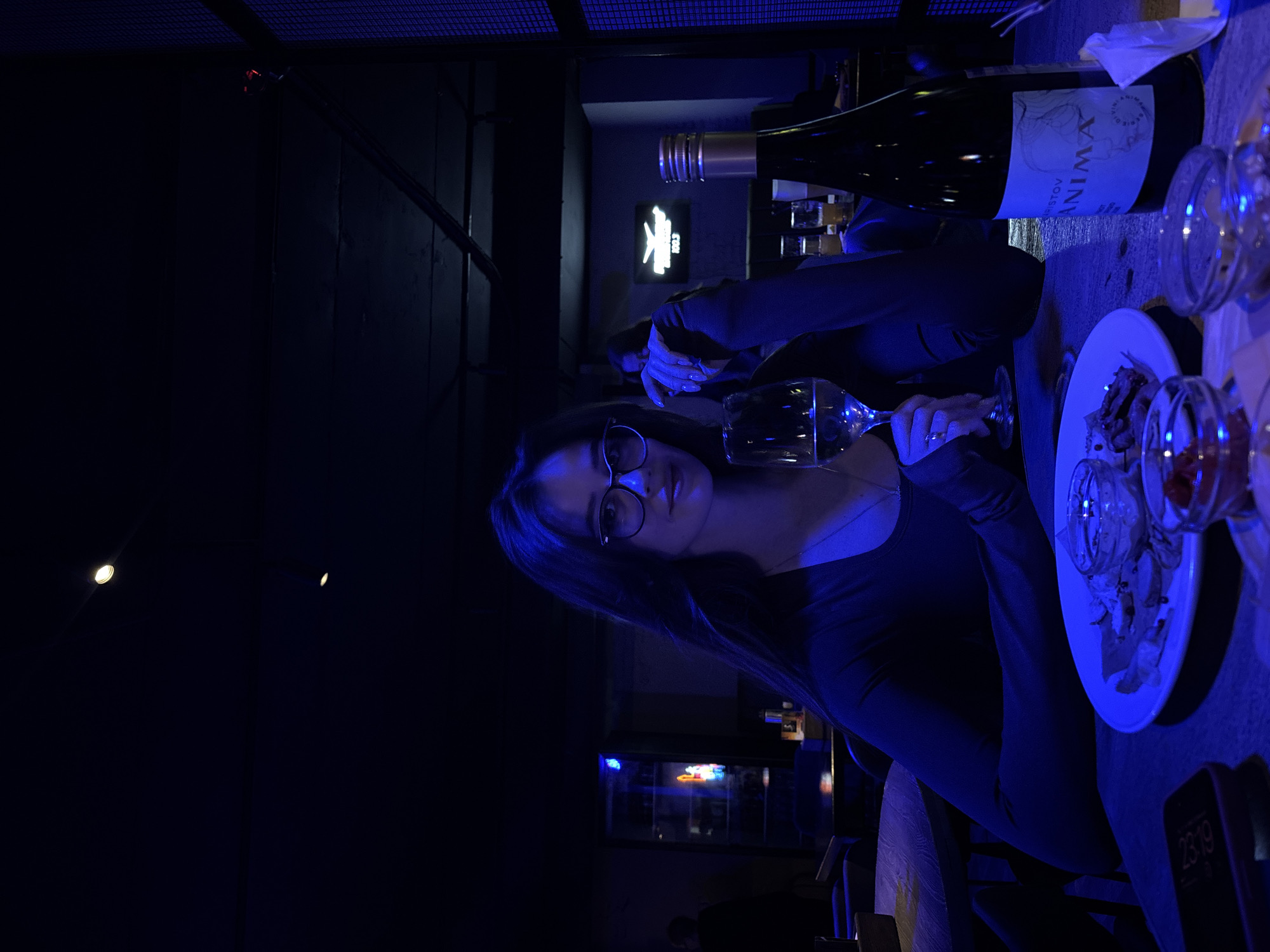 A dimly lit image shows a woman with long dark hair and glasses sitting at a table in what appears to be a bar or restaurant. She is holding a wine glass. On the table is a bottle of wine labeled "ANIMA," a plate with appetizers, and several small glasses. In the background, a neon sign is visible. A mobile phone displaying "23:19" is visible on the right side of the table. The scene has a noticeable blue tint. While the location in the city of Krasnoyarsk, Russia, cannot be confirmed without additional context, the overall setup suggests a casual dining or social setting.