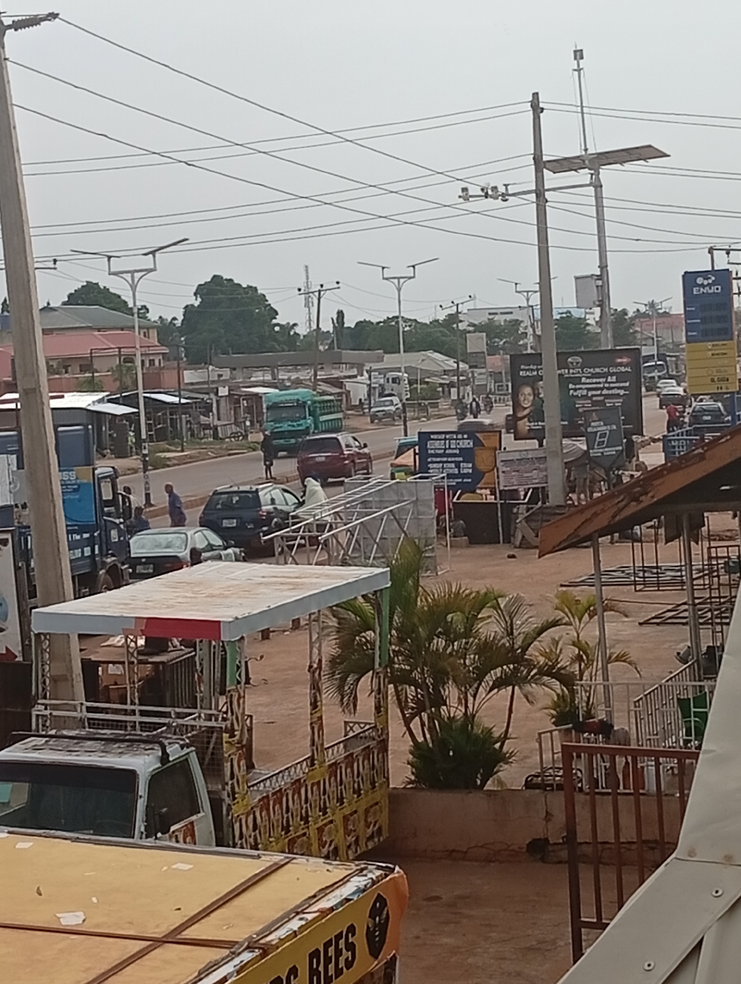 The image depicts a street scene in Sabon Tasha, Nigeria, with a mix of infrastructure and human activity. The view is elevated, looking down onto the street.
Several vehicles are visible, including cars, a truck, and smaller commercial vehicles, some parked and some moving along the road. People are seen walking on the sidewalks and interacting near the vehicles. There are commercial buildings and shops lining the street, with signs and billboards advertising various businesses and services, including an "ENVO" gas station, a church with signs about "Recover All," and another sign for "BEES."
Utility poles with wires and modern streetlights are present, adding to the urban landscape. Vegetation, including small palm trees, is visible in the foreground. Overall, the scene represents a typical, moderately busy commercial area in Sabon Tasha.