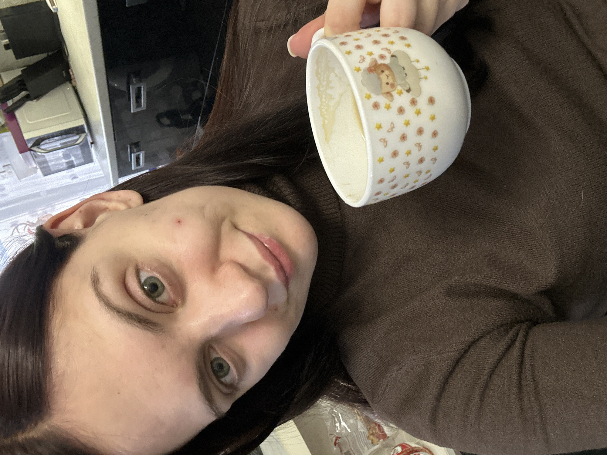A woman with long, dark brown hair and green eyes is holding a white ceramic mug with a sheep and star pattern. The mug appears to contain a small amount of liquid. She is wearing a brown turtleneck sweater. The background shows a kitchen area with a black countertop and cabinets. The location is likely Chaikovsky, Russia.