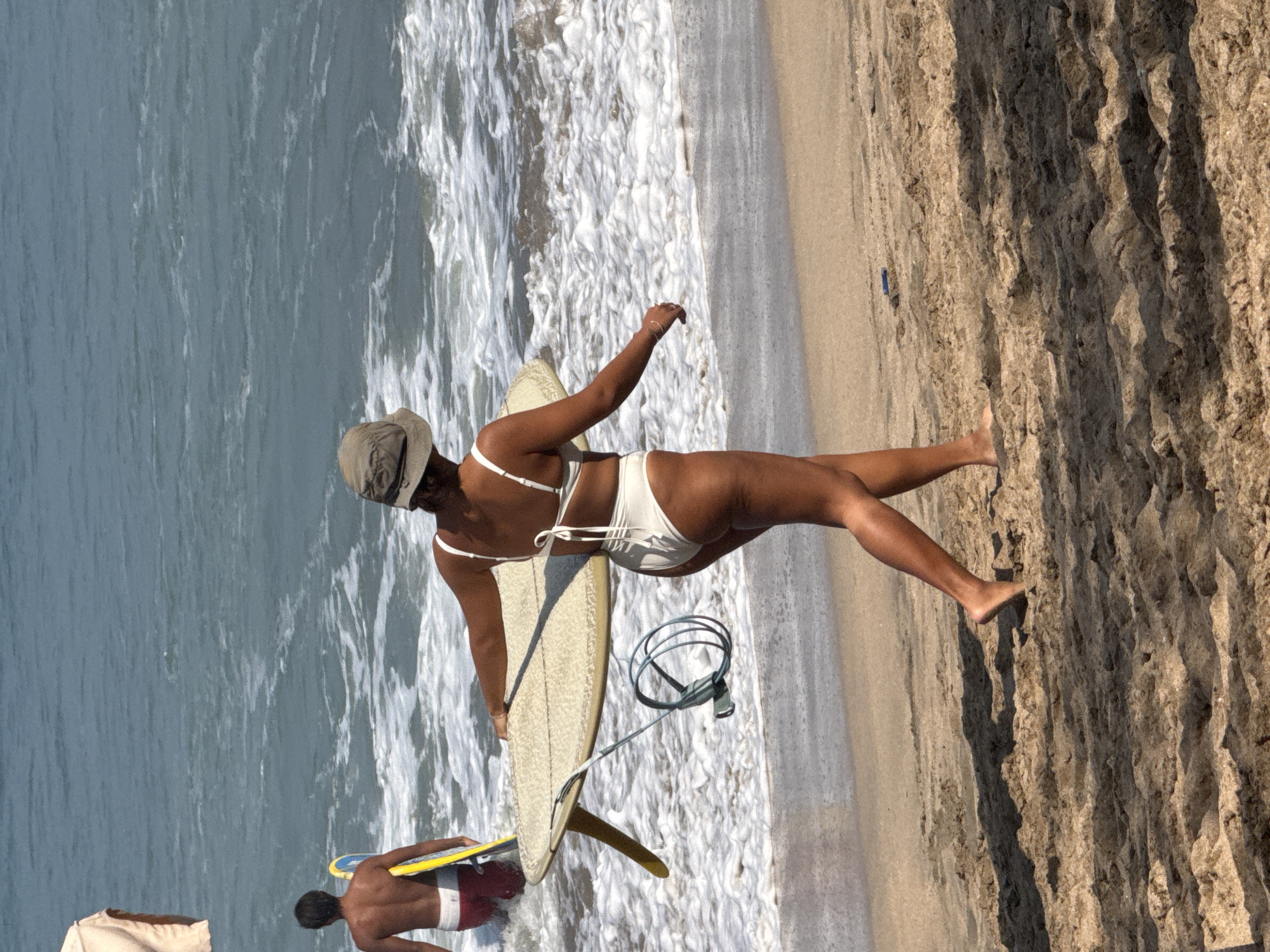 At the beach in Kuta, Indonesia, a woman wearing a white bikini and a light-colored hat walks away from the ocean, carrying a surfboard. Her legs are bare, and she is walking on the sandy shore. Behind her, a man also walks away from the water, holding a yellow surfboard. The ocean waves crash onto the shore, creating white foam. The scene is bathed in sunlight, casting shadows on the sand.