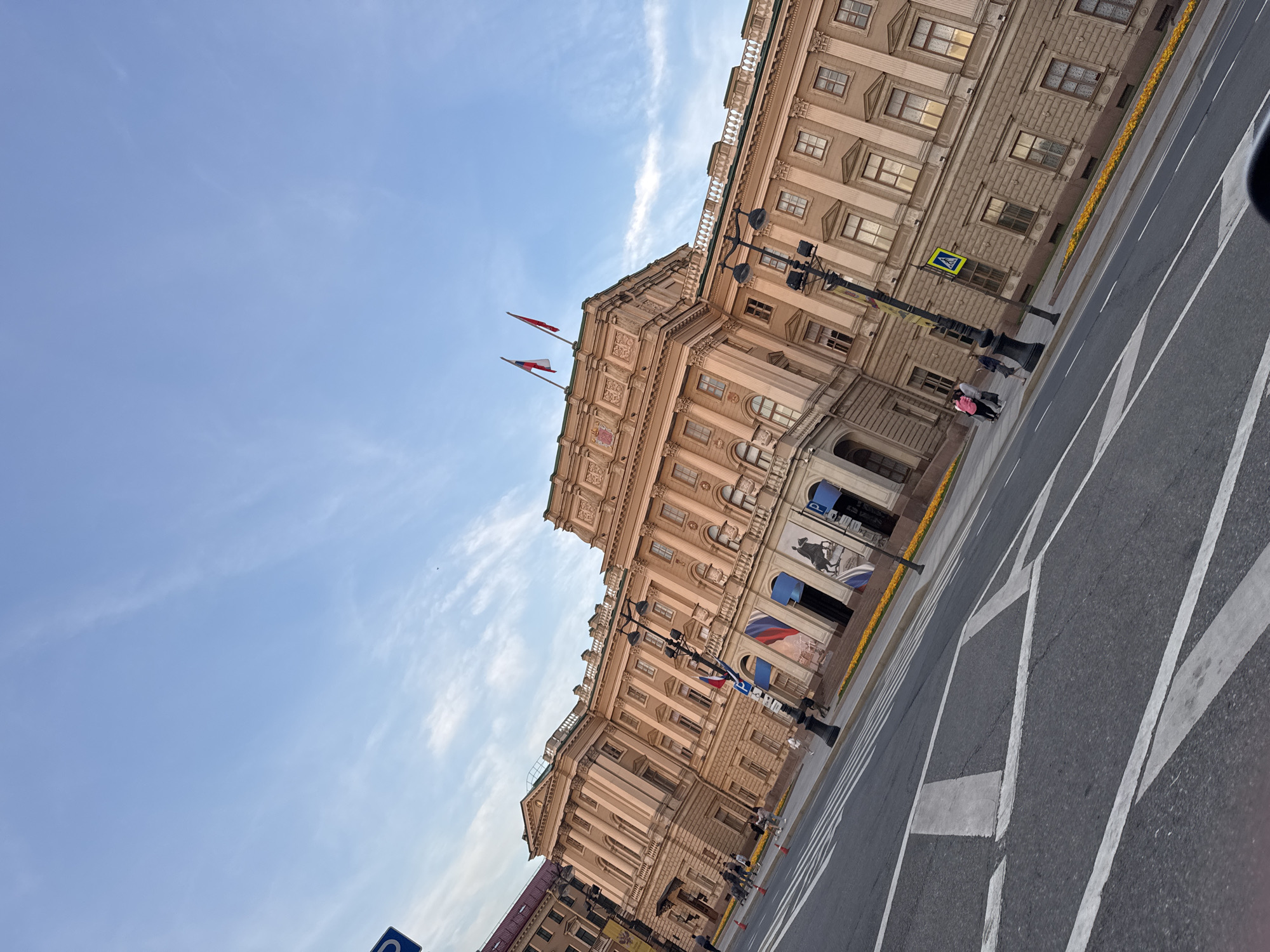 The image captures a street scene in front of a large, ornate building in Saint Petersburg, Russia. The building is a pale beige color with classical architectural features, including columns and decorative moldings. Two flags, one white-red-white and another red-white-blue, fly from the roofline. There are several streetlights and some decorative yellow flowers planted along the building's facade. A pedestrian crossing with white stripes is visible on the asphalt road. A few people are walking on the sidewalk and crossing the street. The sky above is a pale blue with wispy clouds. The perspective of the image is from a slightly elevated, angled viewpoint, suggesting it was taken from a moving vehicle or at an angle. The overall impression is of a grand, historic urban environment. The building is likely the General Staff Building, part of the Hermitage Museum complex on Palace Square.