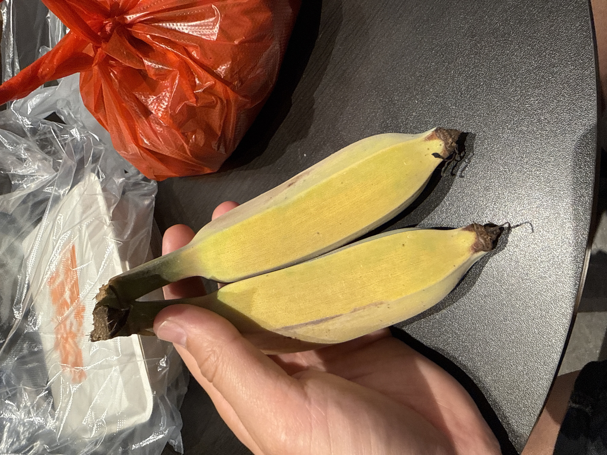 A close-up shot shows a person's hand holding two ripe, yellow bananas. The bananas are connected at the stem and have slightly darkened ends. In the background, there is a translucent plastic bag containing a white foam container and an orange plastic bag. The bananas and the bags rest on a textured, dark gray surface, possibly a table. The image appears to be taken indoors in Liwan District, China.