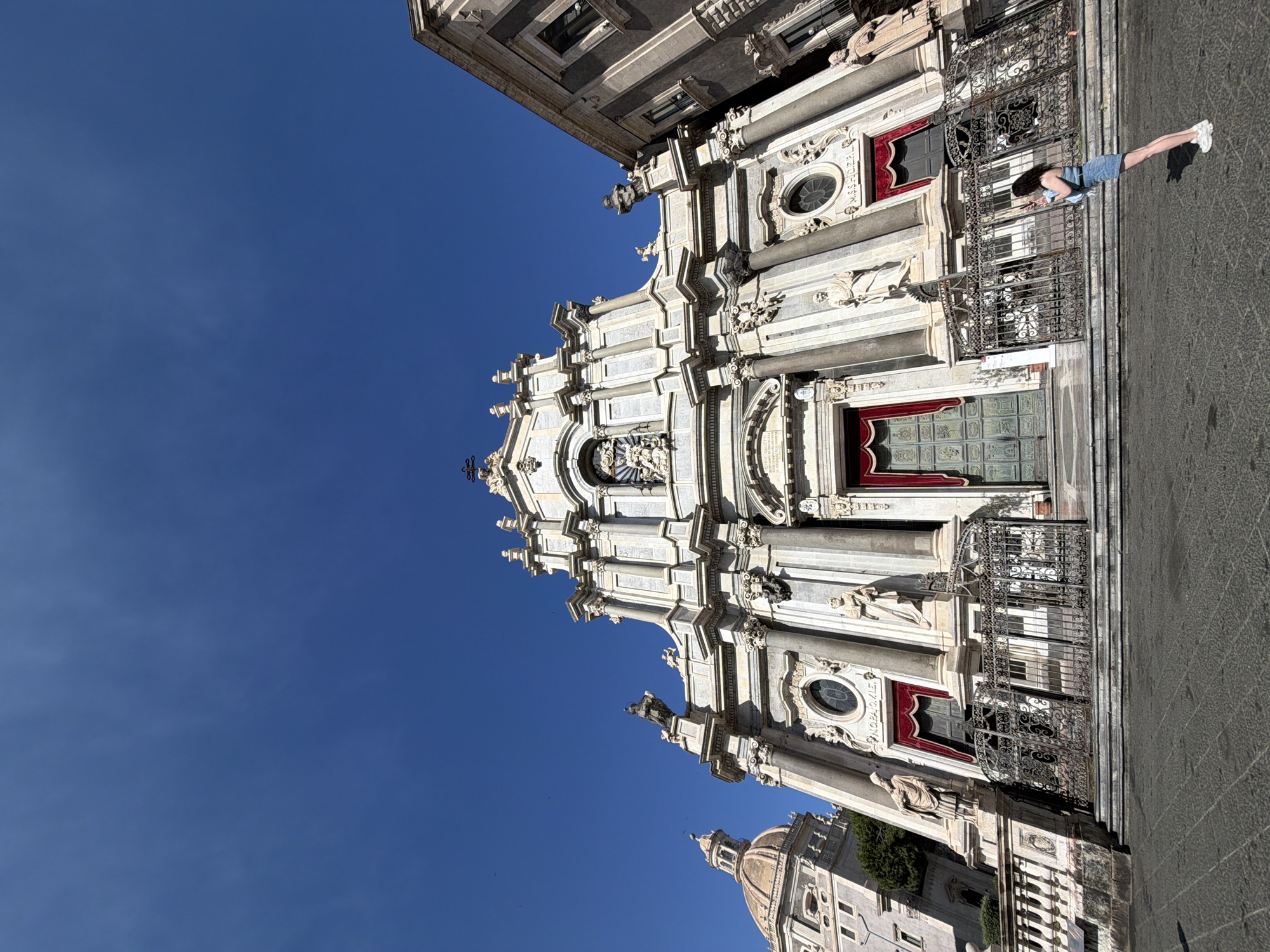 The photograph captures a low-angle, wide shot of the Catania Cathedral (Duomo di Catania) in Catania, Italy. The grand facade of the Baroque-style cathedral dominates the center of the frame, made of white marble and adorned with statues, columns, and intricate carvings. Red velvet curtains hang over the large, green double doors at the entrance. Iron gates are positioned in front of the cathedral steps. To the left of the cathedral, a portion of another building with a dome is visible, also appearing to be made of stone. To the right, a dark-colored building with windows and balconies stands adjacent to the cathedral. In the foreground, a young woman with dark curly hair, wearing a denim skirt and a light-colored top, is standing on the paved plaza, facing away from the camera and looking towards the cathedral. She appears to be taking a photograph with her phone. The sky is a clear, bright blue with no clouds. The lighting suggests it is daytime.