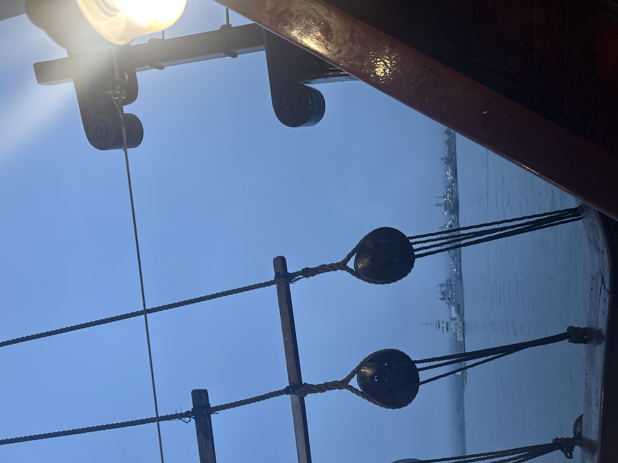 The image shows a view from a sailing ship, likely a replica or historical vessel, in the harbor of Varna, Bulgaria. In the foreground are elements of the ship's rigging: a light fixture, pulleys, and ropes. Through the rigging, one can see the waters of the harbor and, in the distance, the cityscape of Varna. Ships are visible in the harbor. The time appears to be dusk or evening, with a muted, bluish light.