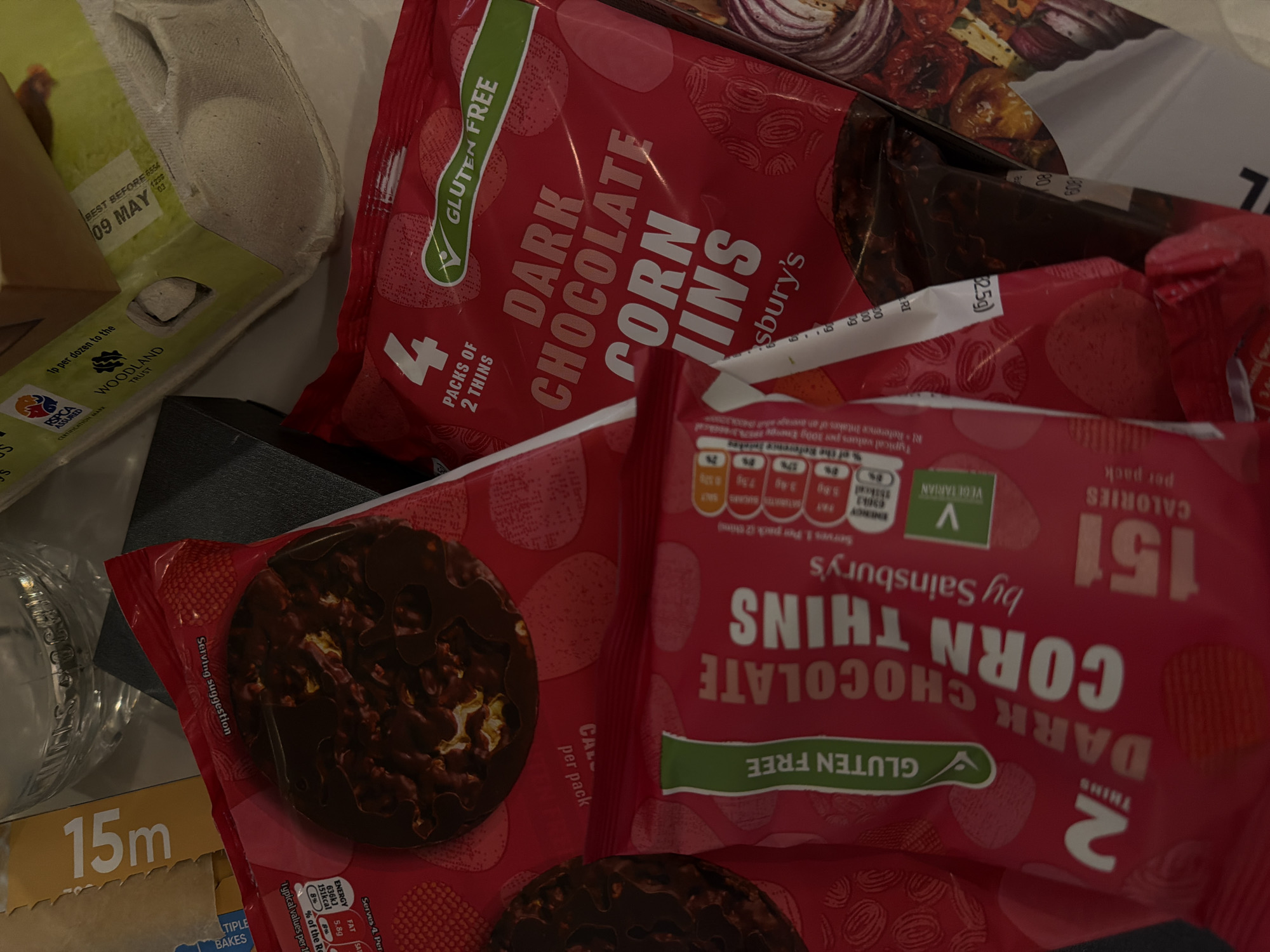 The image displays a collection of grocery items, primarily food products, arranged closely together, possibly on a kitchen surface.
The most prominent items are multiple packages of "DARK CHOCOLATE CORN THINS by Sainsbury's". One large red package in the upper center is labeled "4 PACKS OF 2 THINS" and prominently features "GLUTEN FREE". Another smaller, individual package of the same product is visible in the lower right, displaying "DARK CHOCOLATE CORN THINS", "GLUTEN FREE", and "VEGETARIAN" logos. Nutritional information is visible on this smaller package, indicating "151 CALORIES per pack" and "ENERGY 636kJ 151kcal". A single dark chocolate-coated corn thin is shown as a "Serving suggestion" on one of the packages.
To the upper left, an open carton of eggs is visible, with two white eggs still inside. Text on the carton includes "BEST BEFORE 09 MAY", "1p per dozen to the WOODLAND TRUST", and "RSPCA ASSURED CERTIFICATION MARK". Behind the main red packages, part of another food item's packaging is visible, showing what appears to be sliced red onions or roasted vegetables. In the lower left corner, a brown cardboard box is partially visible, with "15m" and "TIPLE BAKES" printed on it. The scene captures various food products, suggesting recent grocery shopping or items stored together.