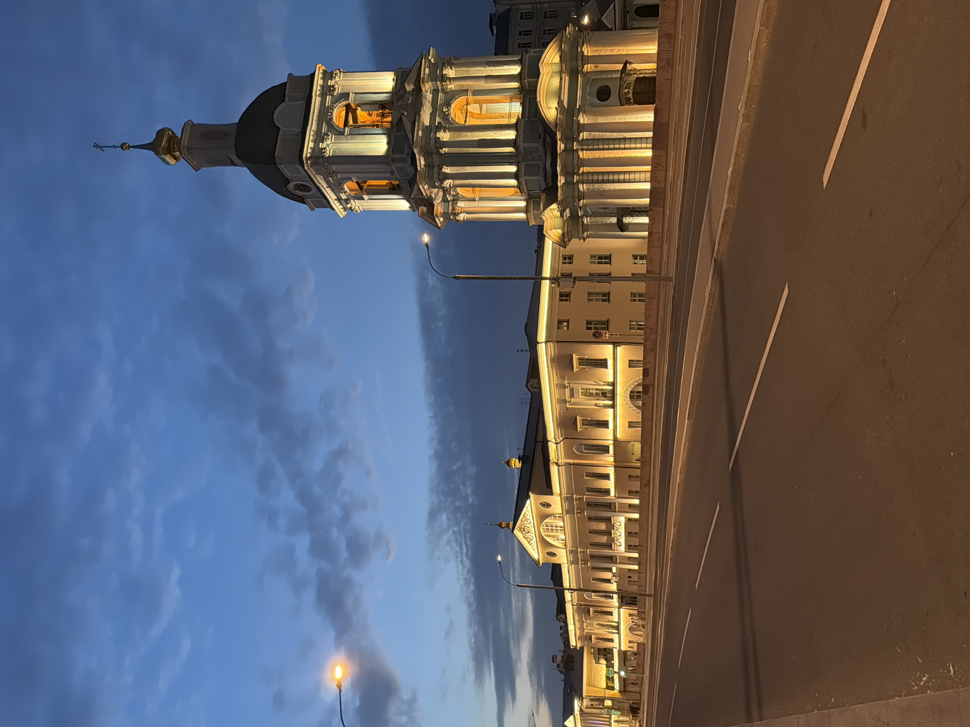 The image depicts a twilight scene in Moscow, Russia, featuring a prominent bell tower of the St. Nicholas Naval Cathedral and a long, illuminated building. The sky is a deep blue with scattered clouds, suggesting the transition from day to night. A street with white lane markings runs in the foreground, indicating a roadway. The bell tower, a white structure with classical architectural elements and a golden dome, is brightly lit, highlighting its intricate details. Adjacent to the tower is a long, neoclassical building, also illuminated, with numerous windows and decorative features. Streetlights are visible, adding to the ambiance. No people are visible in the frame. The overall impression is one of architectural grandeur and tranquil evening illumination.