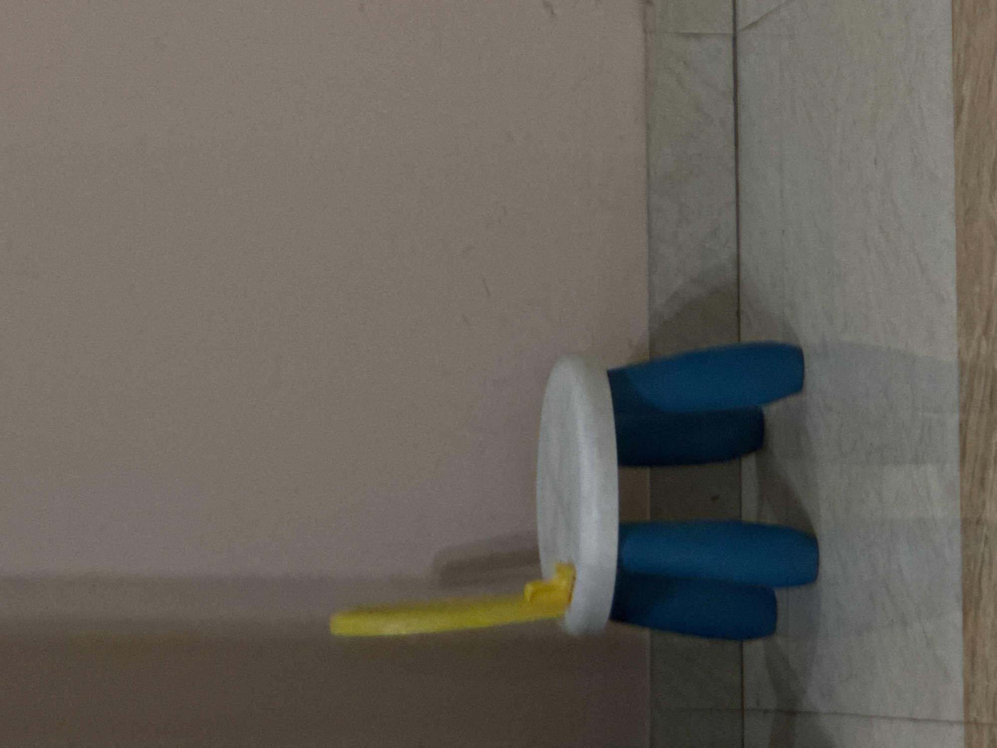The image shows a small, child-sized chair against a wall and floor. The chair has a white circular back, blue legs, and a yellow seat. The environment consists of a plain wall and tiled flooring. The photo does not provide any information to determine that the location is in Yerevan, Armenia.