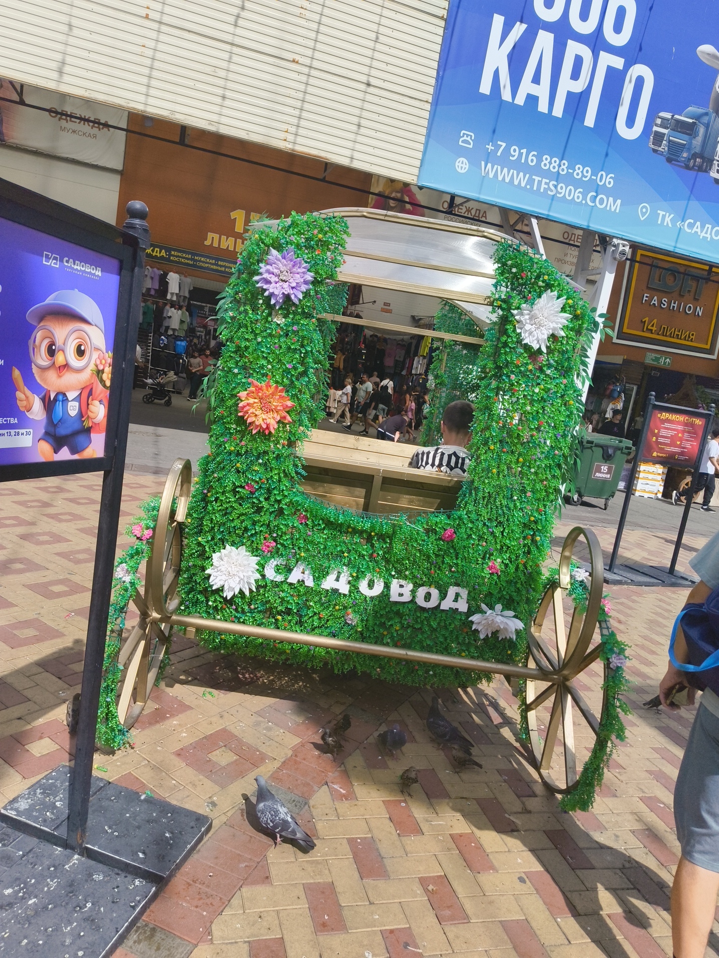 This image depicts a decorative, green, carriage-like structure covered in artificial foliage and adorned with large artificial flowers. The words "САДОВОД" (Sadovod) are spelled out in white letters on the side of the structure. The carriage has two decorative wheels and appears to be a prop or exhibit. In the background, there are shops and a banner for "LOFT FASHION" and "14 ЛИНИЯ" (14 Line), suggesting a shopping area. Another banner in the upper left advertises "ОДЕЖДА МУЖСКАЯ" (Men's Clothing). A digital display featuring an owl character is also visible on the left. Several pigeons are present on the paved ground in the foreground. The scene suggests an outdoor marketplace or shopping center, likely in Russia given the Cyrillic text. A young person is seated inside the carriage, facing away from the camera. The overall setting is a bright, sunny day.