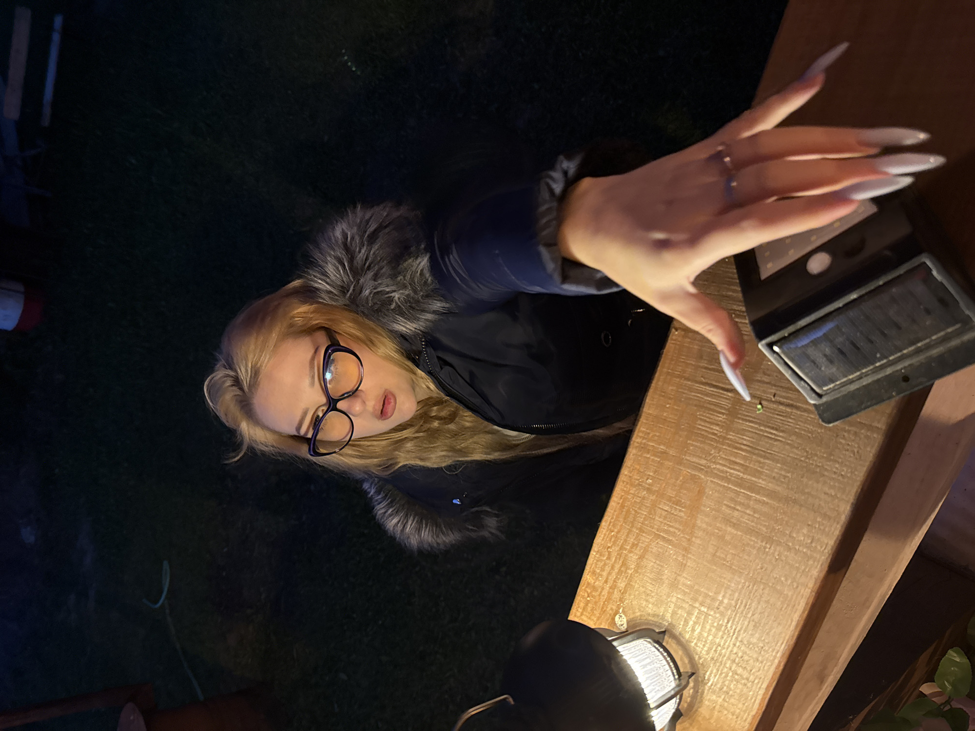 A young woman with blonde hair and glasses is reaching out her hand towards the camera. She is wearing a dark winter coat with a fur-lined hood. The background is dark and appears to be an outdoor setting, possibly a backyard, with grass and some wooden structures visible. A lit lamp and a solar-powered light are on a wooden surface in the foreground. The overall lighting suggests it is nighttime. The woman's hand has long, pointed fingernails, and she is wearing a ring. There are no explicit indications of the specific location within Большая Венья, Russia, beyond it being an outdoor, potentially residential area.