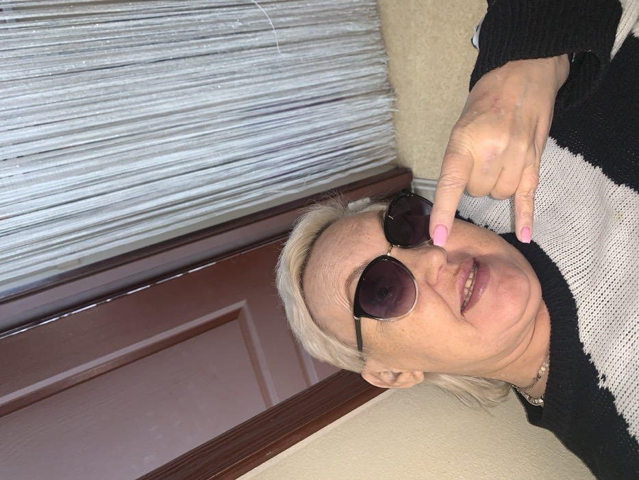 This image depicts an adult female positioned indoors, stated to be in Chaikovsky, Russia. The subject has short, light-colored hair and is wearing black and gold-rimmed sunglasses with dark lenses. She is dressed in a black and white striped or color-blocked knitted sweater and a light-colored beaded necklace. Her fingernails are painted with light pink polish. The woman is looking directly towards the camera with a slight smile, lips parted. Her right hand is raised, performing a "sign of the horns" gesture with her index finger and pinky extended, positioned close to her nose. The background features a light-colored wall to the left, and a dark brown wooden door frame and door panel in the upper left and center. To the right, a vertical string or bead curtain of numerous light-colored strands is visible, with a light-colored floor surface appearing in the lower right. The image is captured from a low, slightly upward angle.