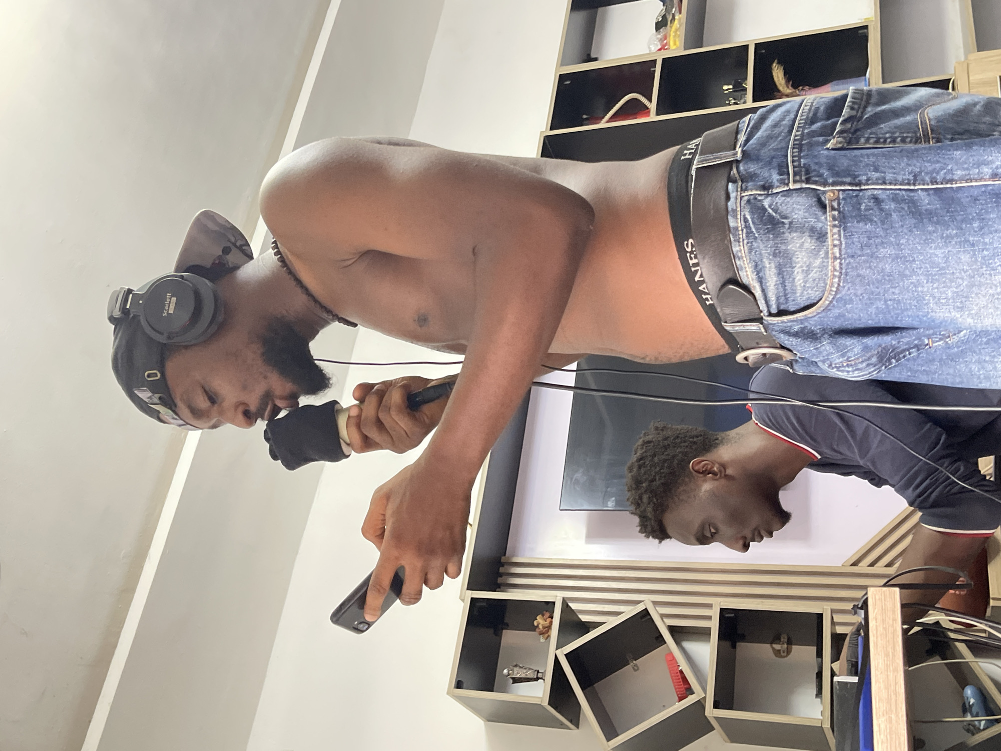 A shirtless, dark-skinned man with a beard, wearing jeans, a black Hanes underwear waistband, headphones, and a headscarf, is holding a microphone wrapped in black fabric in his left hand and a cell phone in his right hand. He is standing in what appears to be an indoor space with a wall-mounted shelving unit behind him. The location is identified as Abuja, Nigeria. Another person is visible in the reflection in the television. They are wearing a dark blue shirt with white and red details.