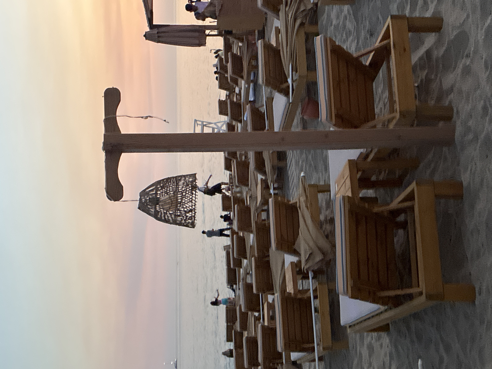 The image depicts a beach scene at dusk in Alanya, Turkey. In the foreground, several empty wooden lounge chairs are arranged on the sand. A wooden post with a woven hanging lamp is prominent in the center of the frame. In the midground, a few people are visible near the water's edge, some walking and others standing. The sea is calm, and a faint line of a sailboat can be seen on the horizon. The sky displays a soft gradient of pastel colors, indicating the sunset. The overall atmosphere is peaceful and relaxed.