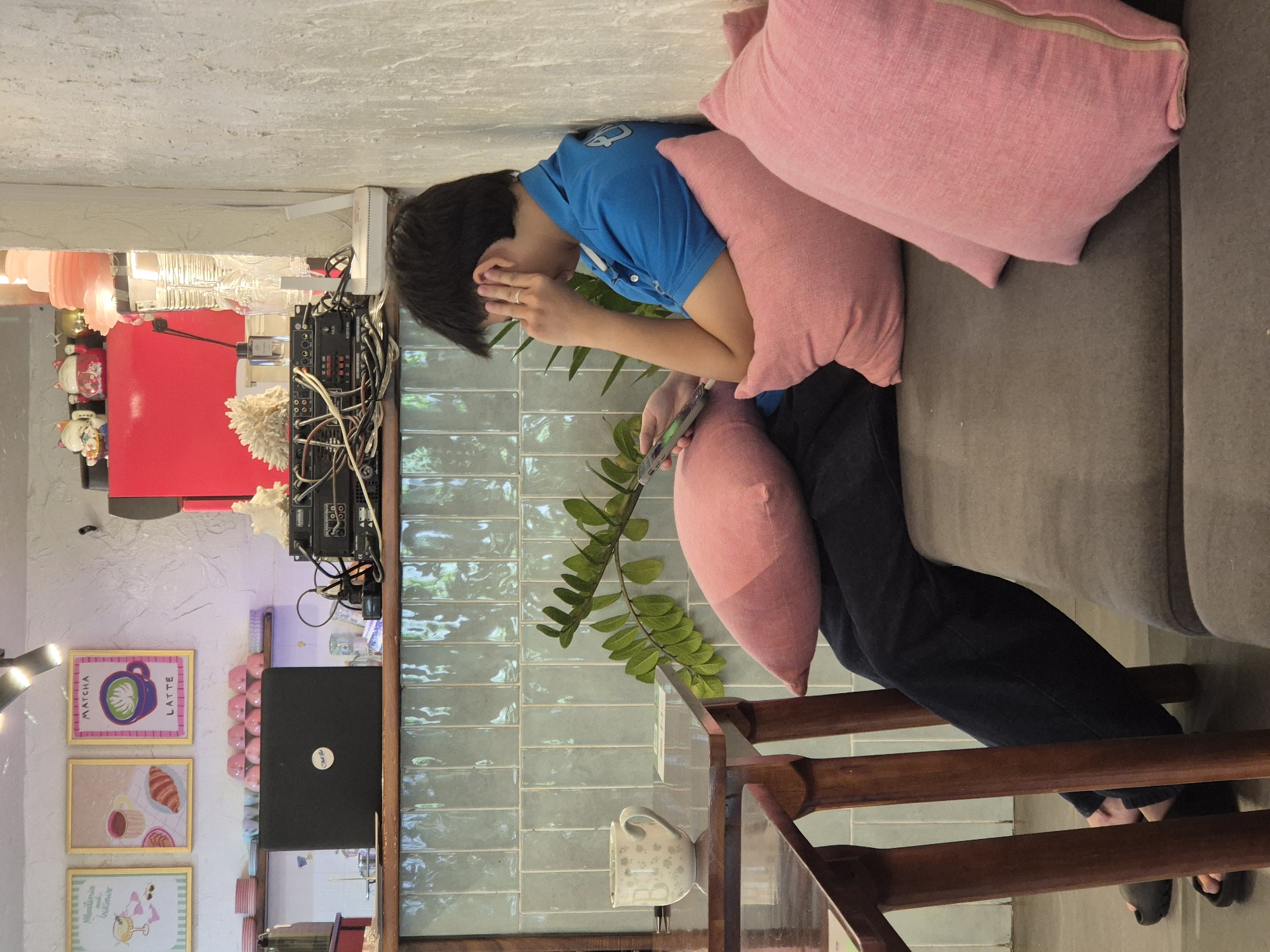 A young person sits on a couch in a cafe in Da Nang, Vietnam, looking at their phone and holding their ear. The person is wearing a blue polo shirt and dark pants. Pink cushions are on the couch and behind the person. A table with a cup and a plant is to the left. In the background, there are framed pictures on the wall, a laptop, and audio equipment.